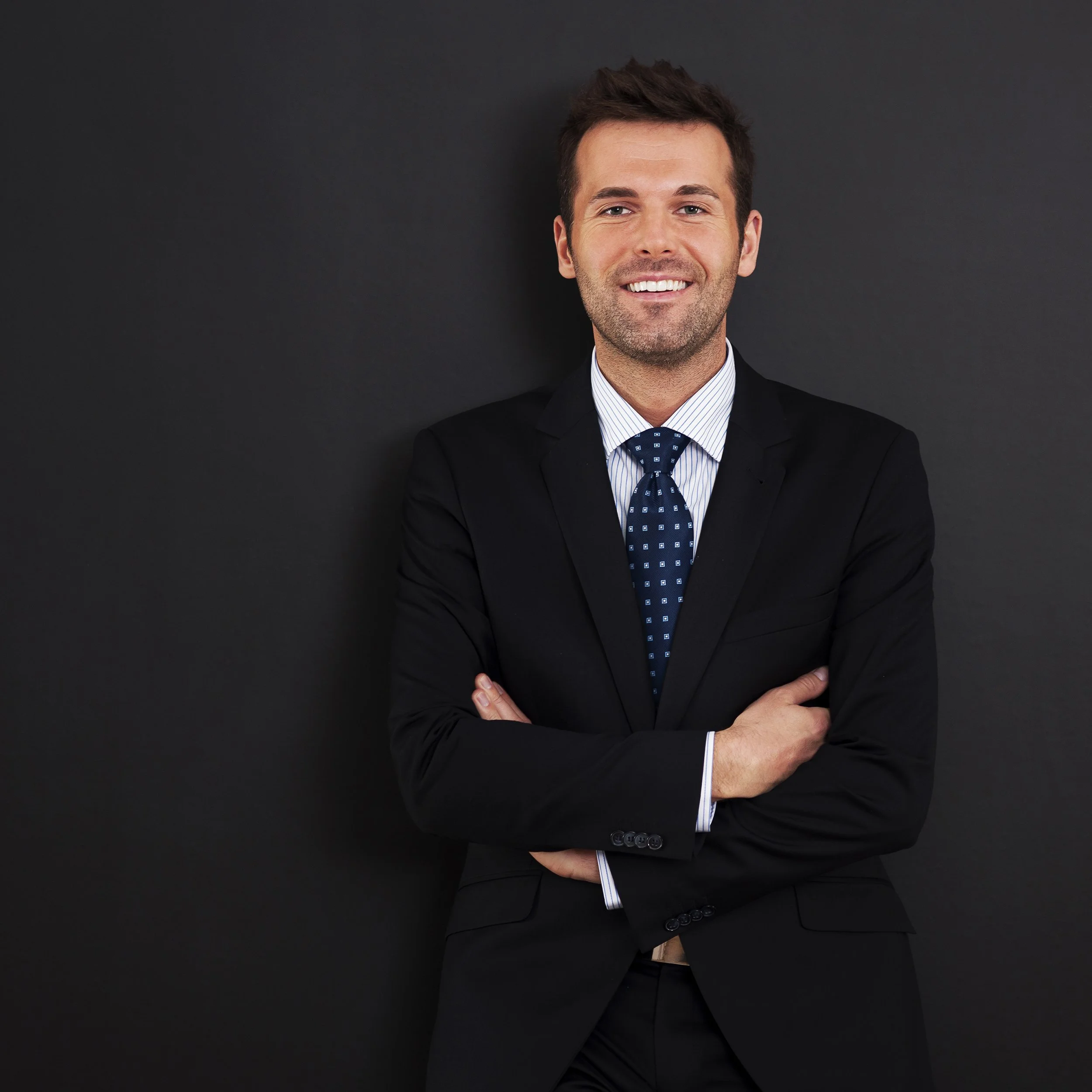 A man dressed in a black suit with a white shirt and patterned blue tie, standing against a dark background with his arms crossed and smiling.