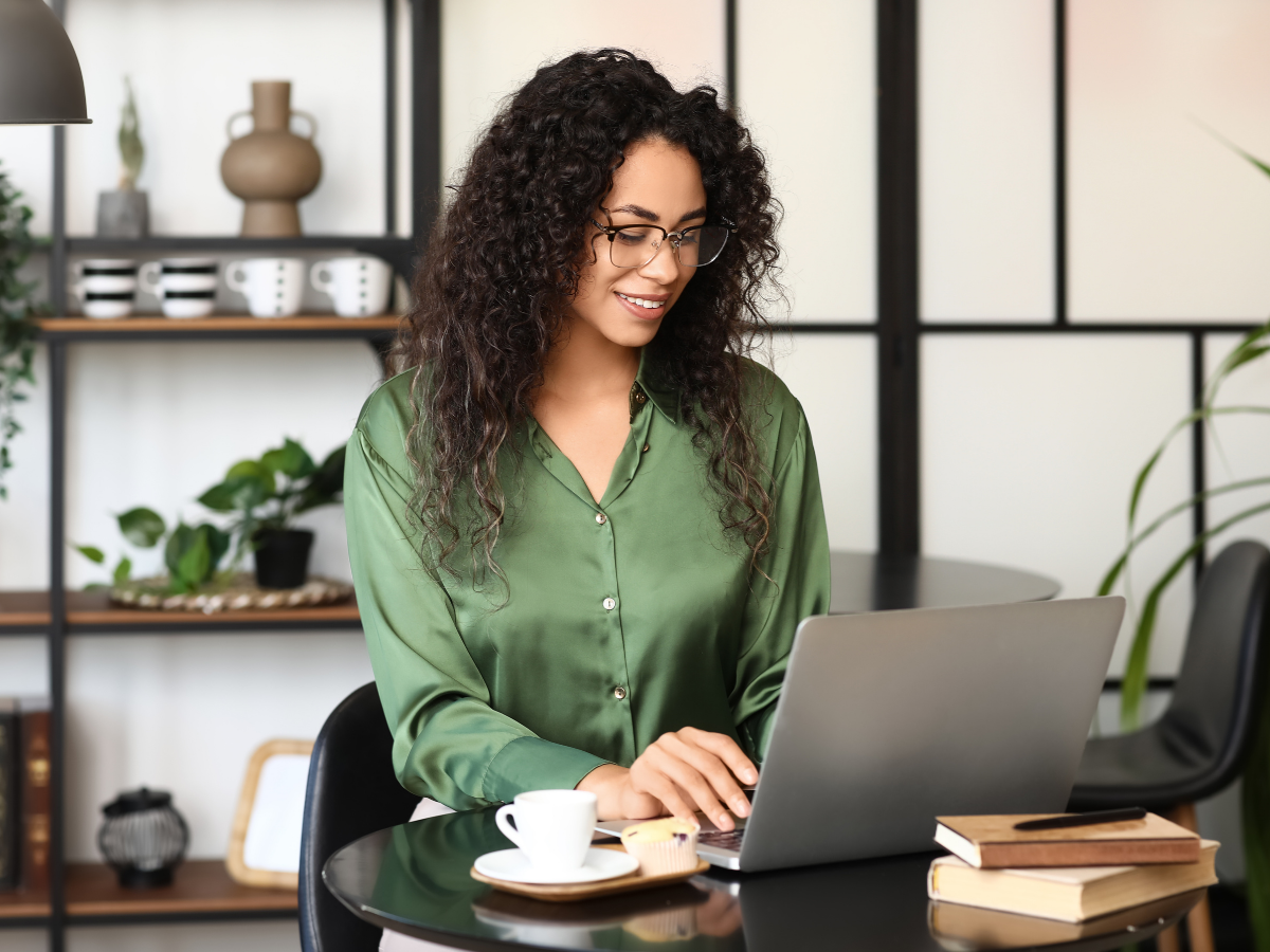 Woman with curly hair and glasses working on a laptop in a modern, decorated office with plants and shelves.