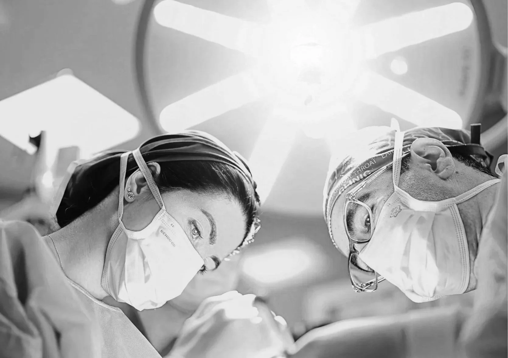 Two surgeons wearing masks, goggles, and scrubs perform a surgery under bright operating room lights. The image is taken from an angle looking up at the surgical team.