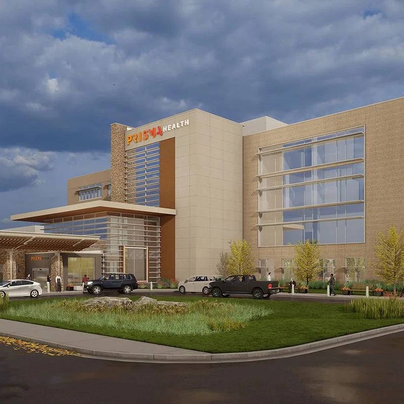 Modern hospital building with the sign 'PRISMA HEALTH' at the top, a driveway with parked cars, a few trees, and a partly cloudy sky.