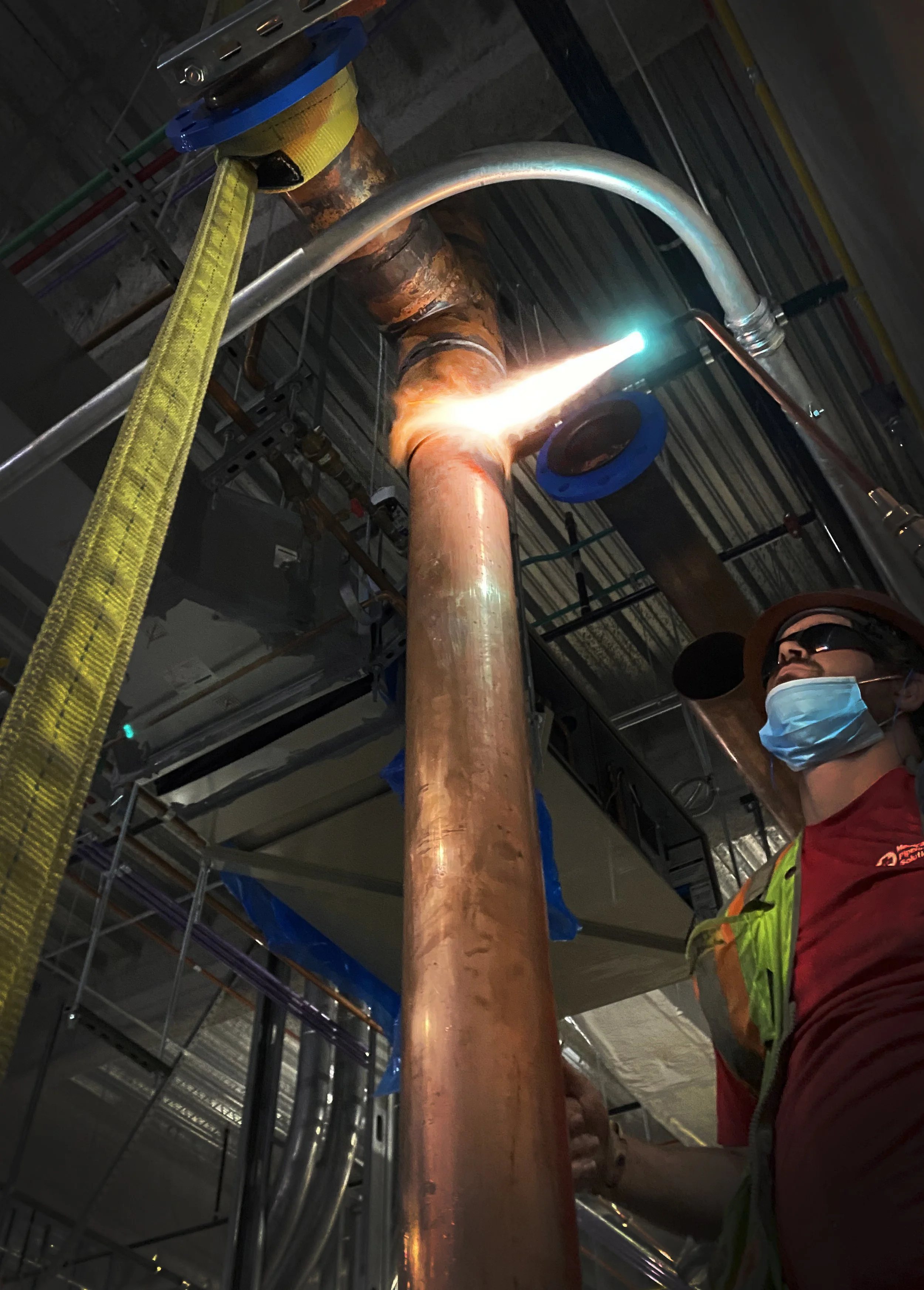 Worker in safety gear, including mask and sunglasses, brazing a large copper pipe in an industrial setting, with sparks and bright light from the brazing process.