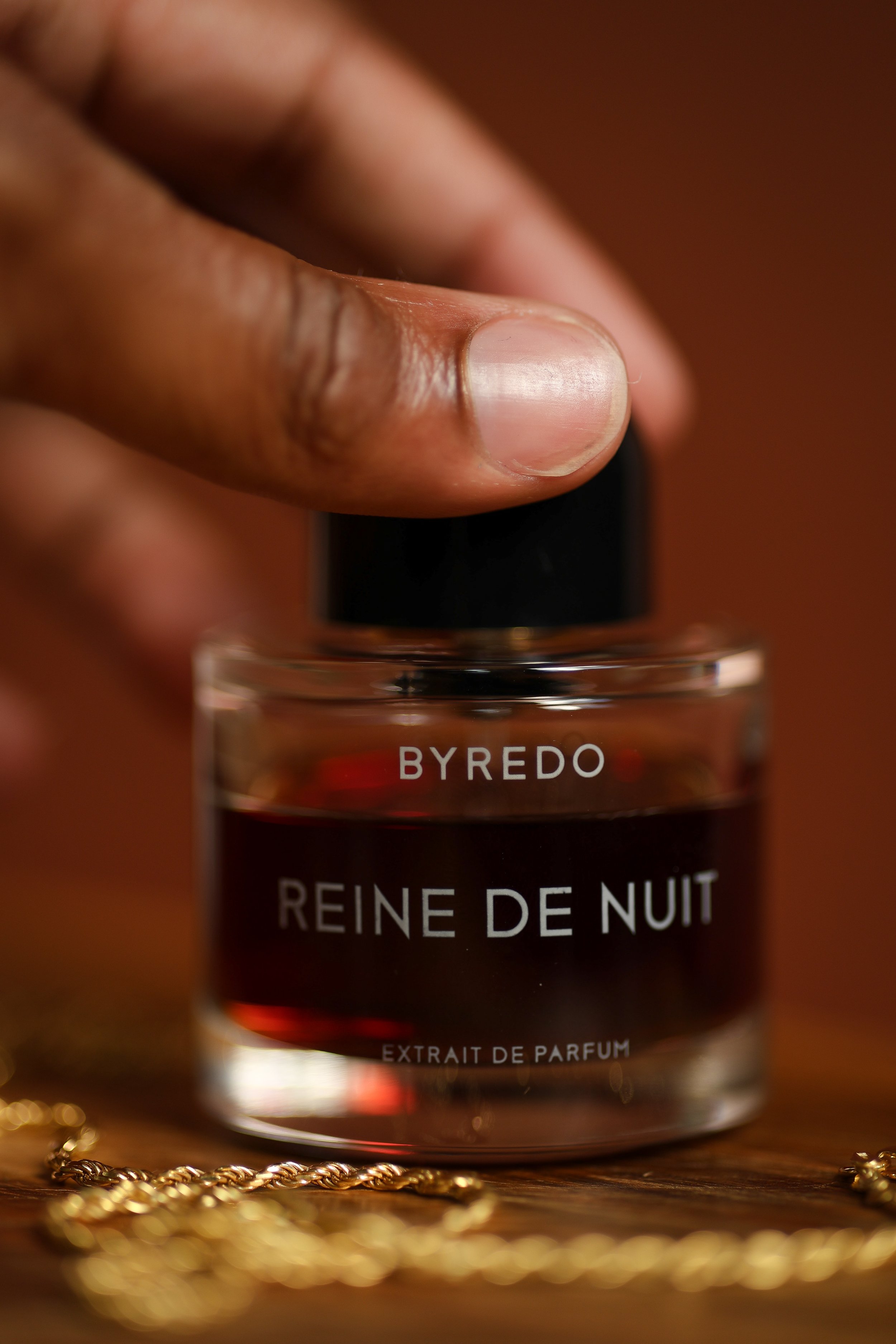 Close-up of a hand holding a small perfume bottle labeled 'Reine de Nuit' by Byredo, with a gold chain in the foreground.