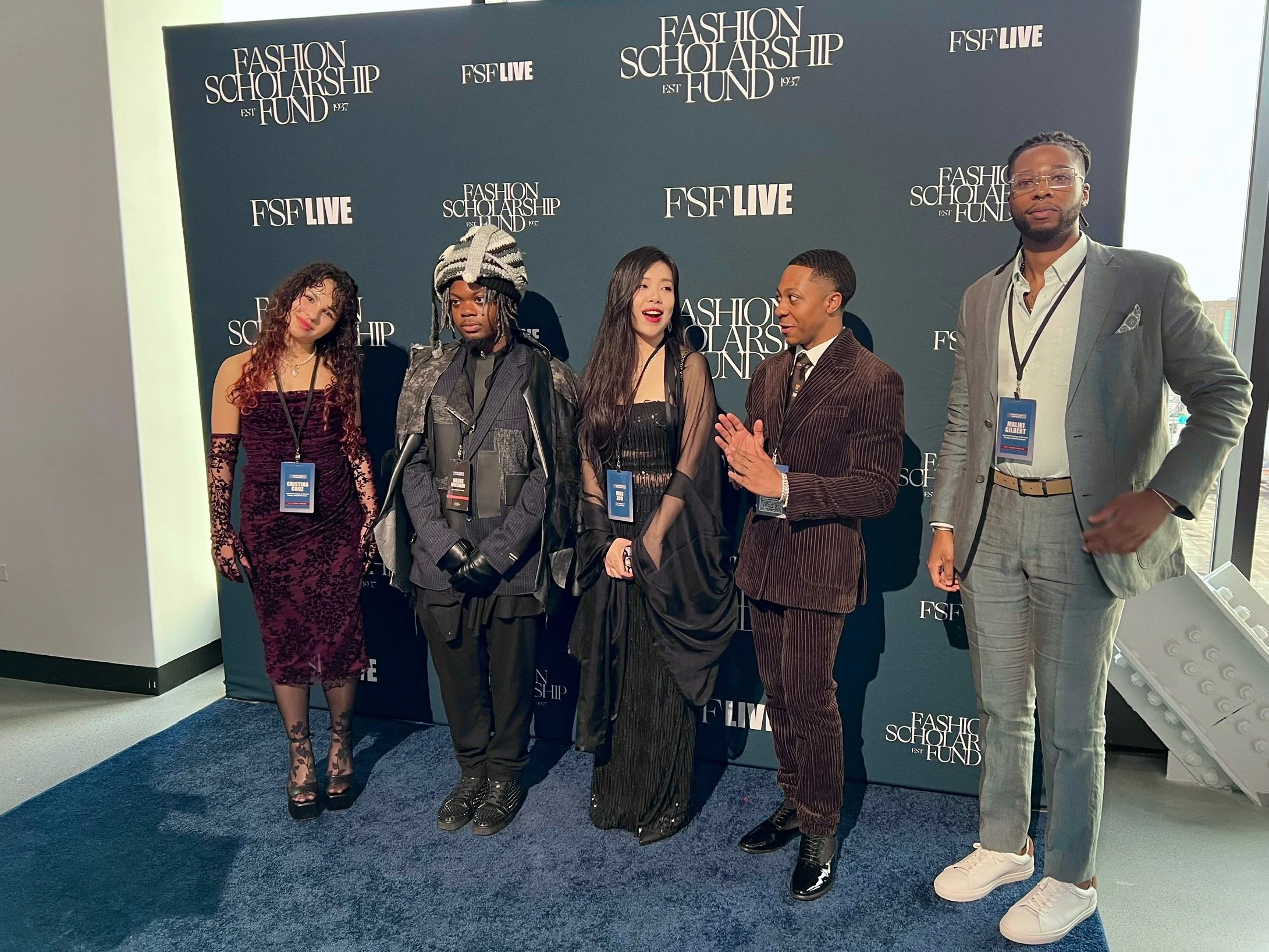 Group of five diverse individuals dressed in formal attire standing in front of a dark blue backdrop with white text that reads 'Fashion Scholarship Fund' and 'FSF Live.' They are at an event, posing for a photo.