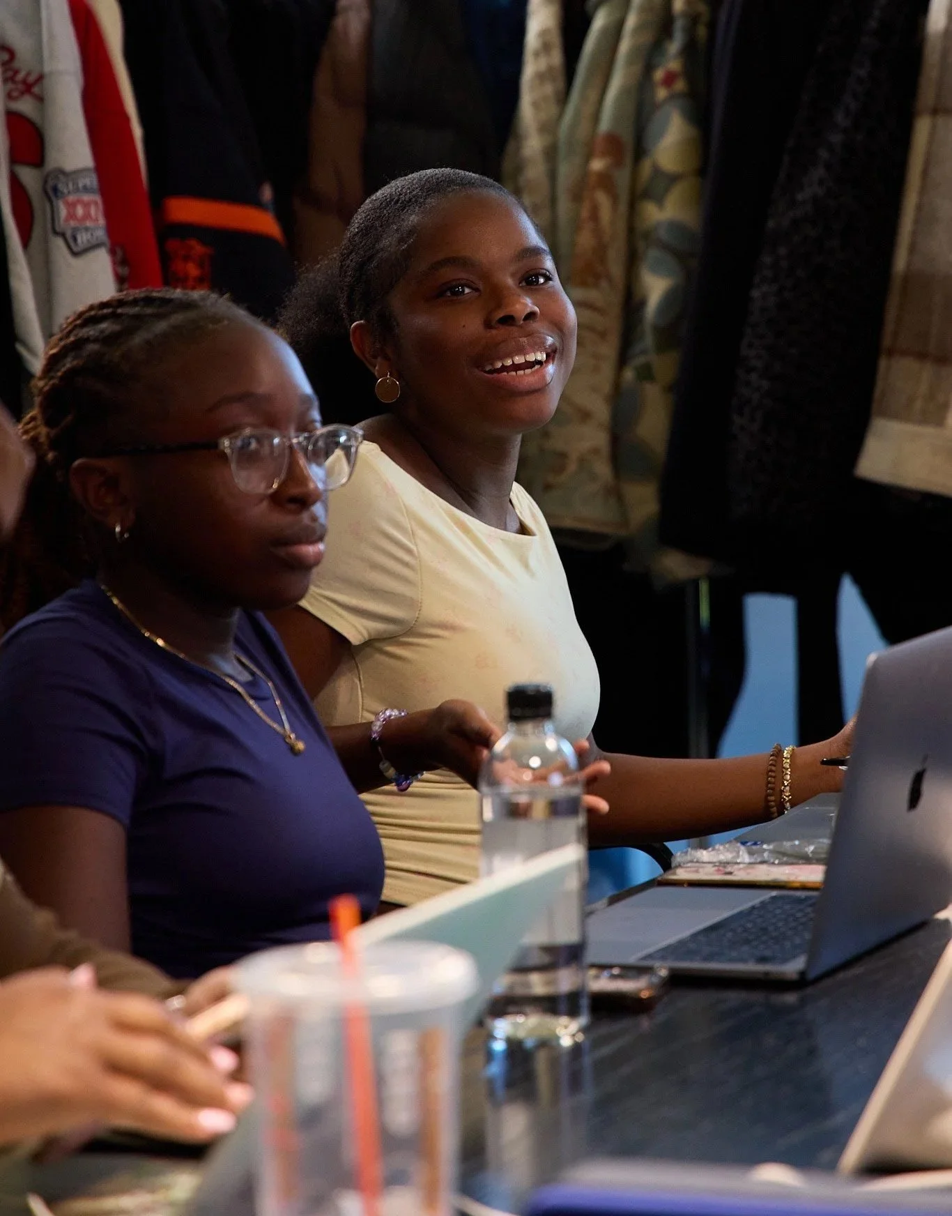 Two women sitting at a table with laptops and drinks, engaged in a conversation or presentation, in an indoor setting.
