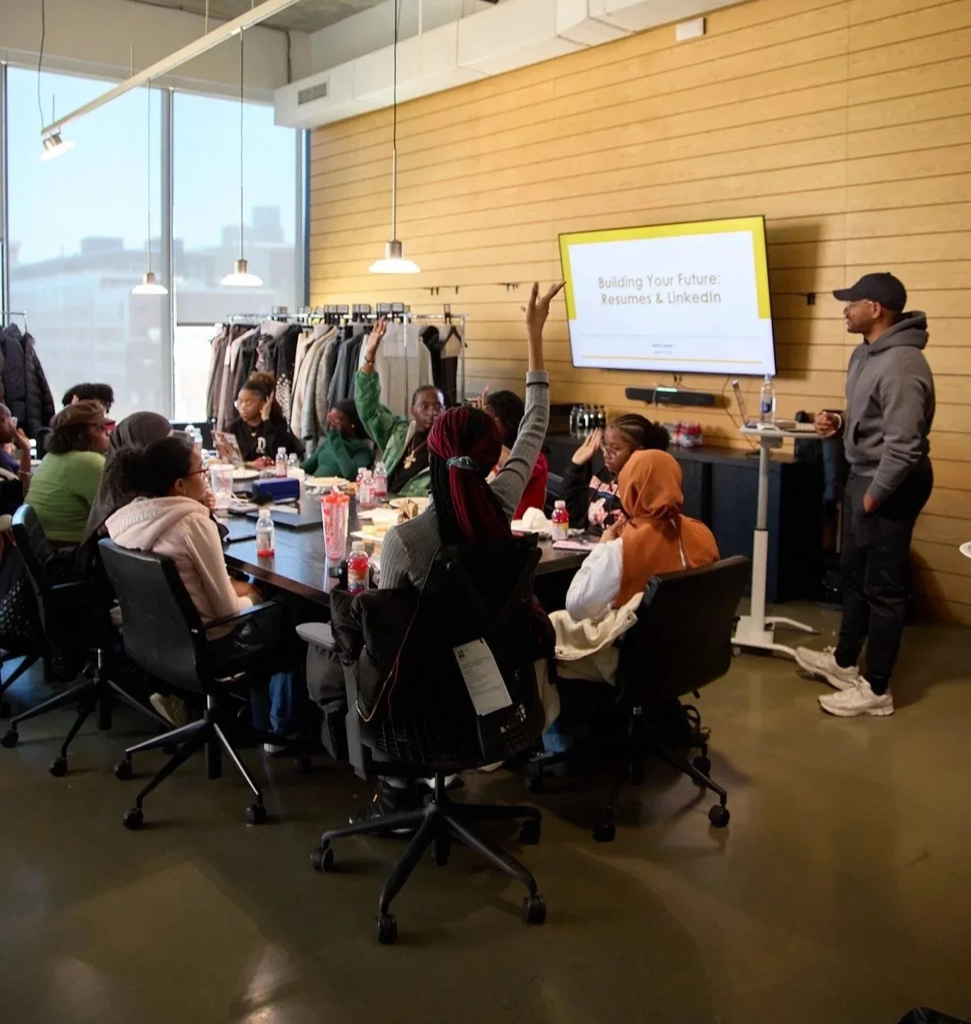 A group of people attending a seminar in a room with large windows. Some attendees are raising their hands. A presenter stands near a screen displaying the title 'Building Your Future: Resumes & LinkedIn.' Clothing racks with jackets are visible in t