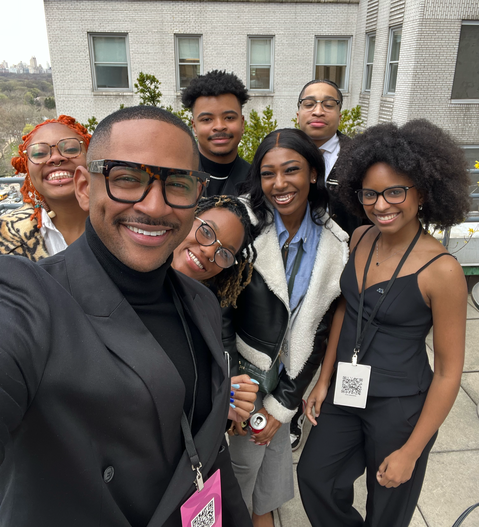 A group of seven diverse young adults smiling and taking a selfie outdoors on a rooftop terrace with a cityscape in the background.
