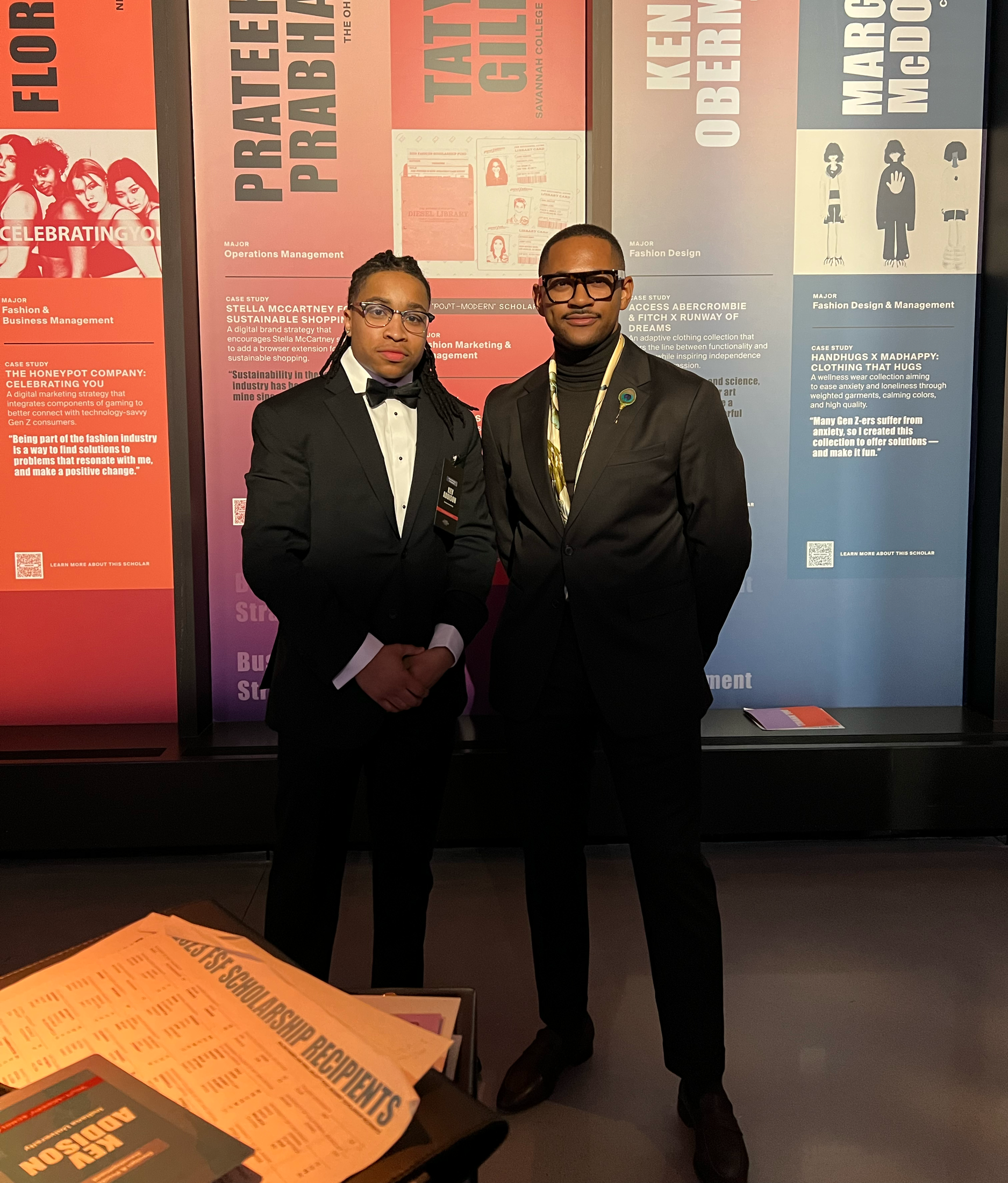 Two men in formal attire standing in front of a display board with research summaries, one wearing glasses and a tuxedo with a bow tie, the other wearing a suit with glasses and a lanyard, at an academic or professional event.