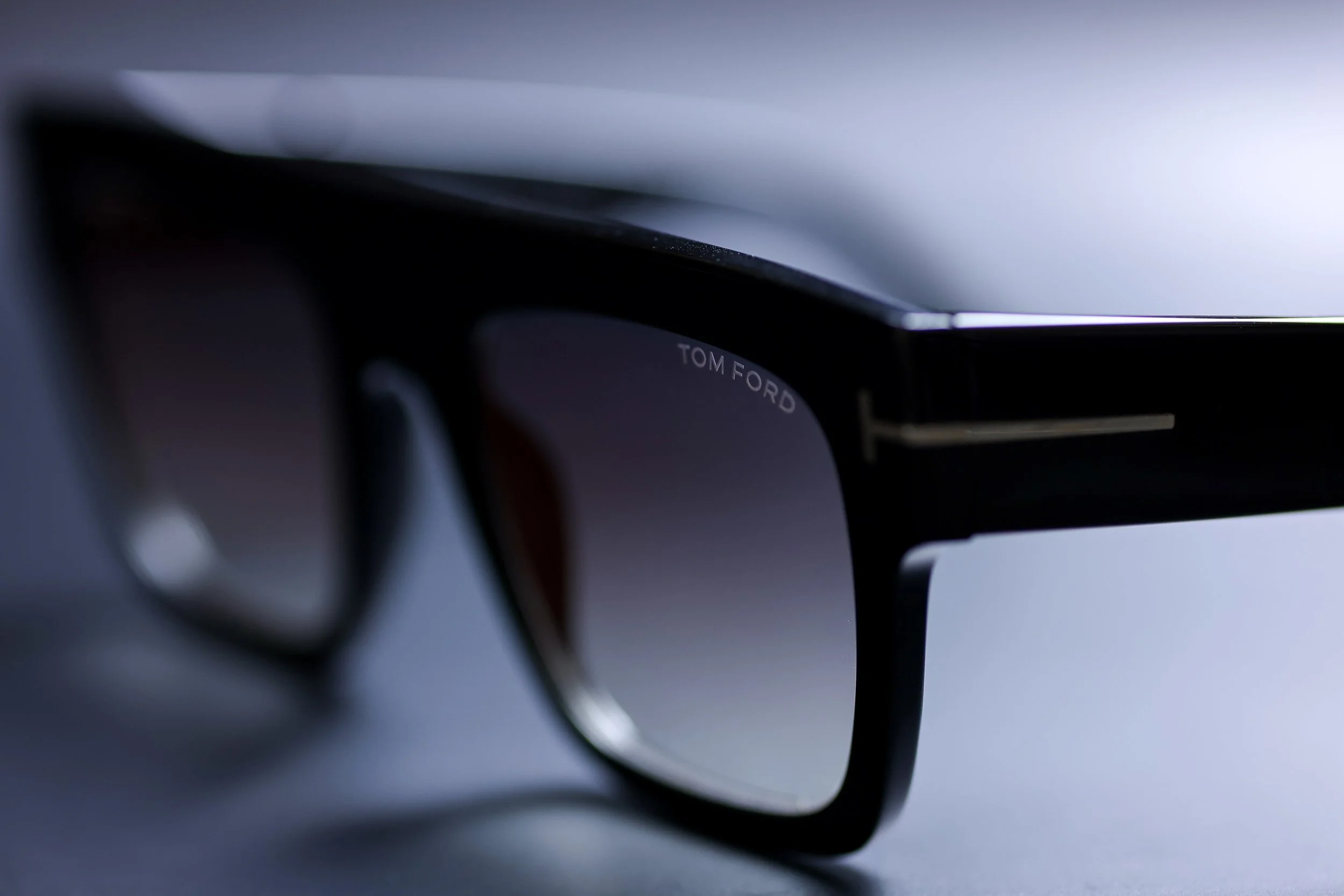 Close-up of black sunglasses with "TOM FORD" branding on the temple, resting on a flat surface with a blurred background.