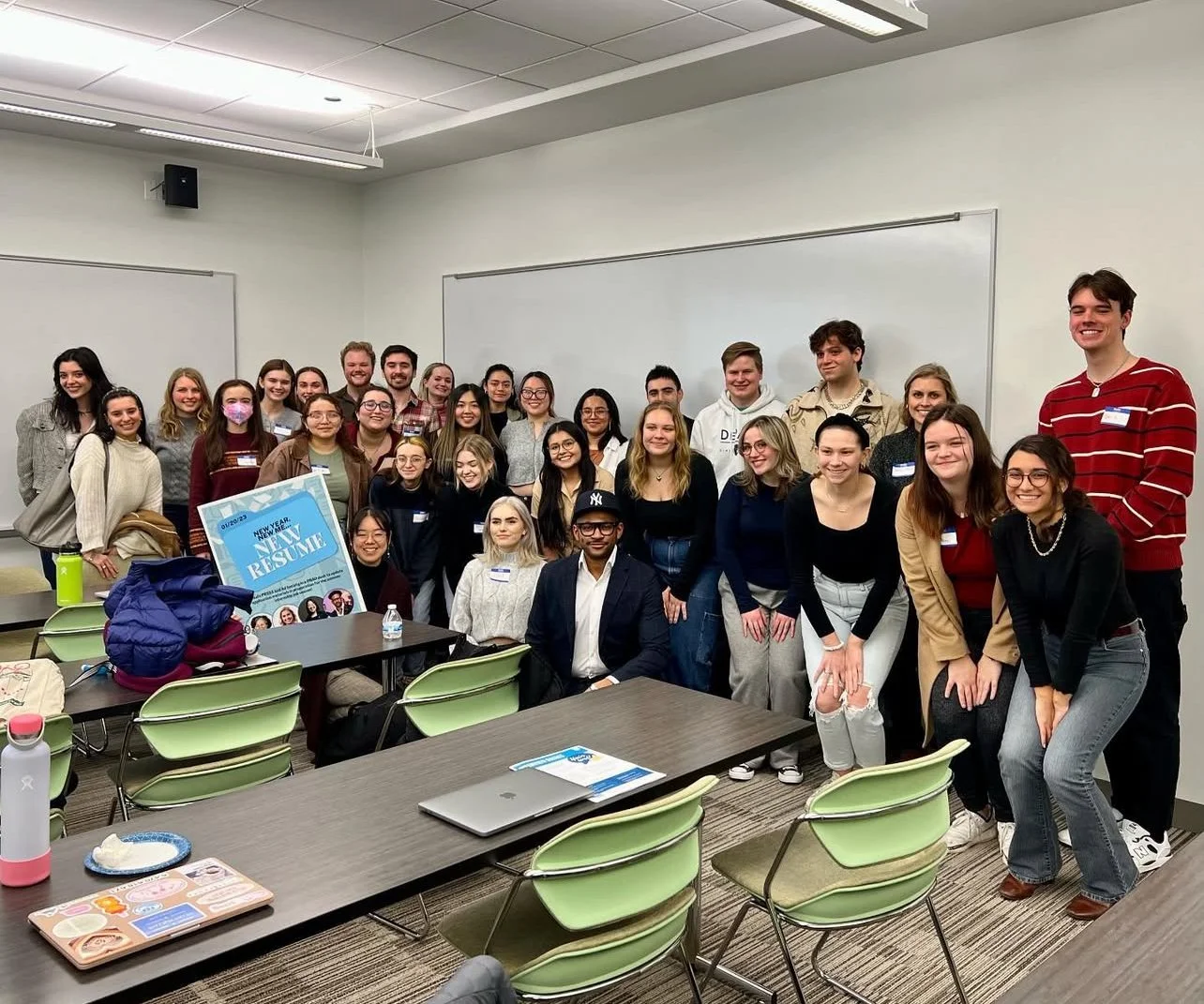 Group of young adults gathered in a classroom, posing for a photo. There are tables and chairs, some with personal items and a résumé poster.