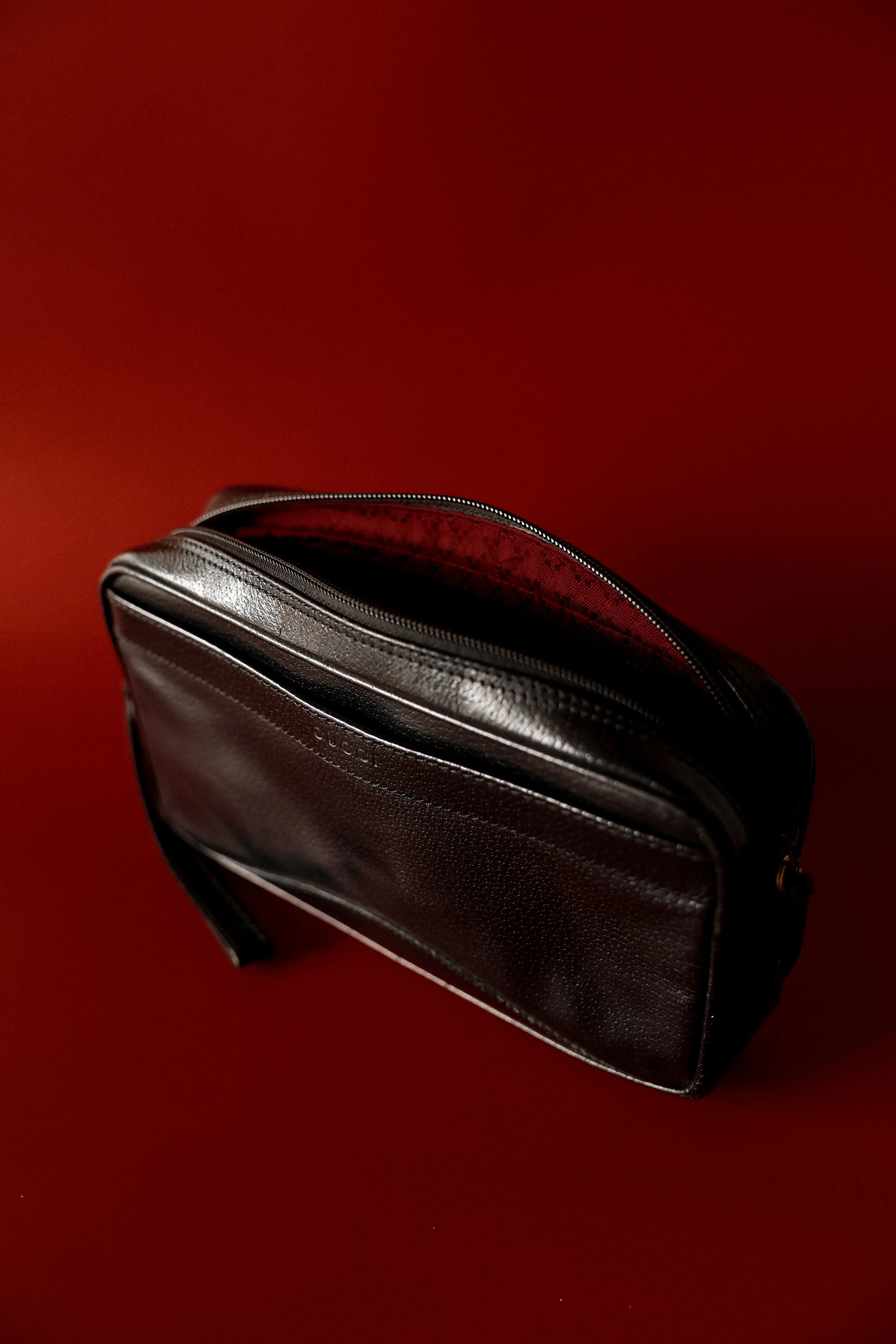 A black leather shoulder bag with a zipper, placed on a red surface. The bag has a front pocket with subtle branding and an inner red lining visible through the unzipped top.