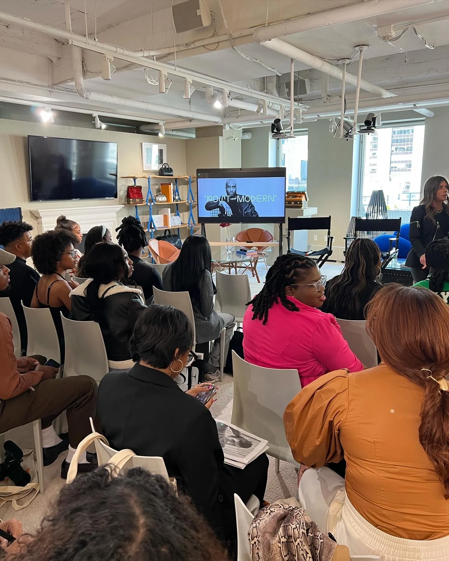 A group of people seated in a modern conference room attending a presentation. There is a woman standing to the right, and a large screen at the front displaying the words 'Root Modern.' The room has natural light coming in through windows and contem