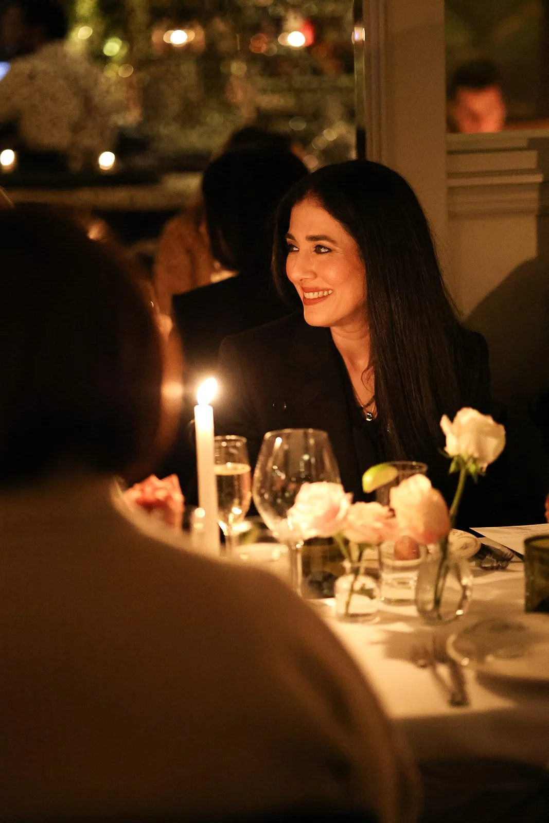 A woman with long dark hair and a bright smile sitting at a dinner table with flowers, wine glasses, and a lit candle in a warmly lit restaurant or home setting.