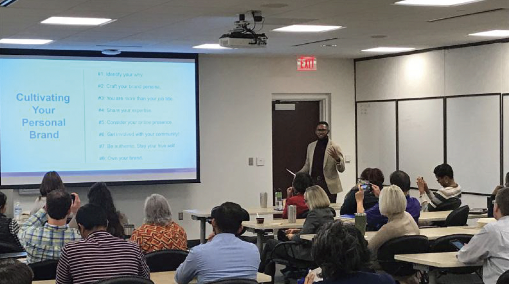 A presenter stands in front of a classroom, giving a presentation on cultivating a personal brand. The slide lists goals such as identifying your why, crafting your brand persona, and owning your brand. The audience sits at desks, some taking notes o