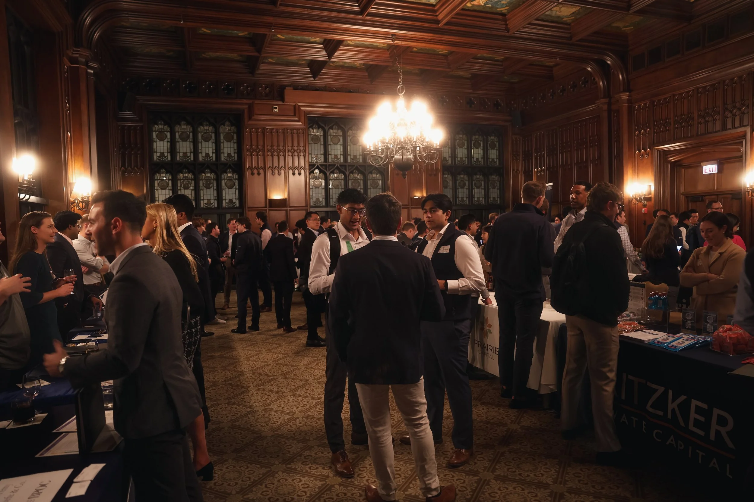 People networking and mingling at a formal event in a grand, wood-paneled room with stained glass windows and a large chandelier overhead.