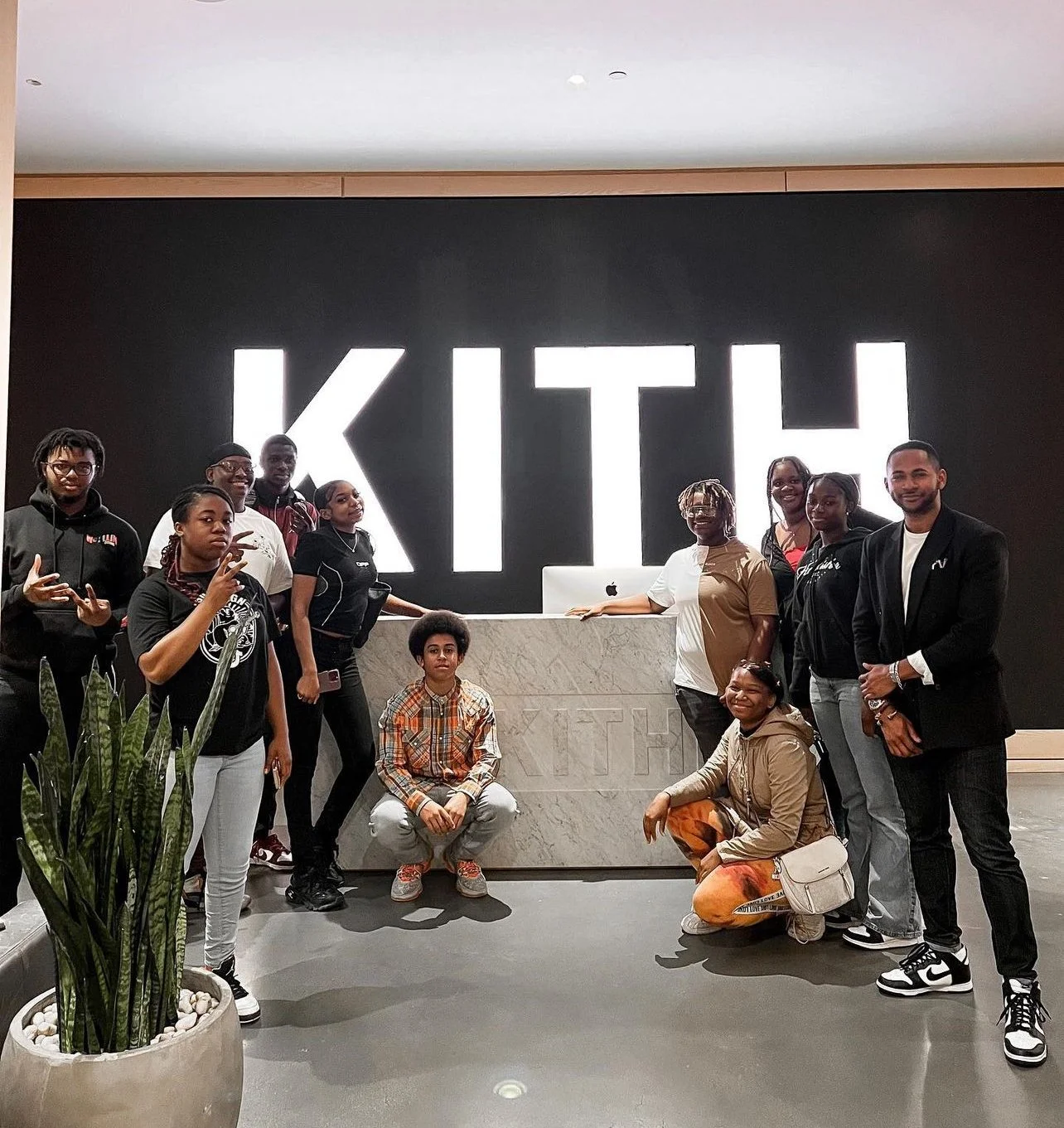 Group of ten people posing in front of a large illuminated sign spelling 'KITH' at a modern office or lobby reception area, with a potted plant in the foreground and a marble reception desk.