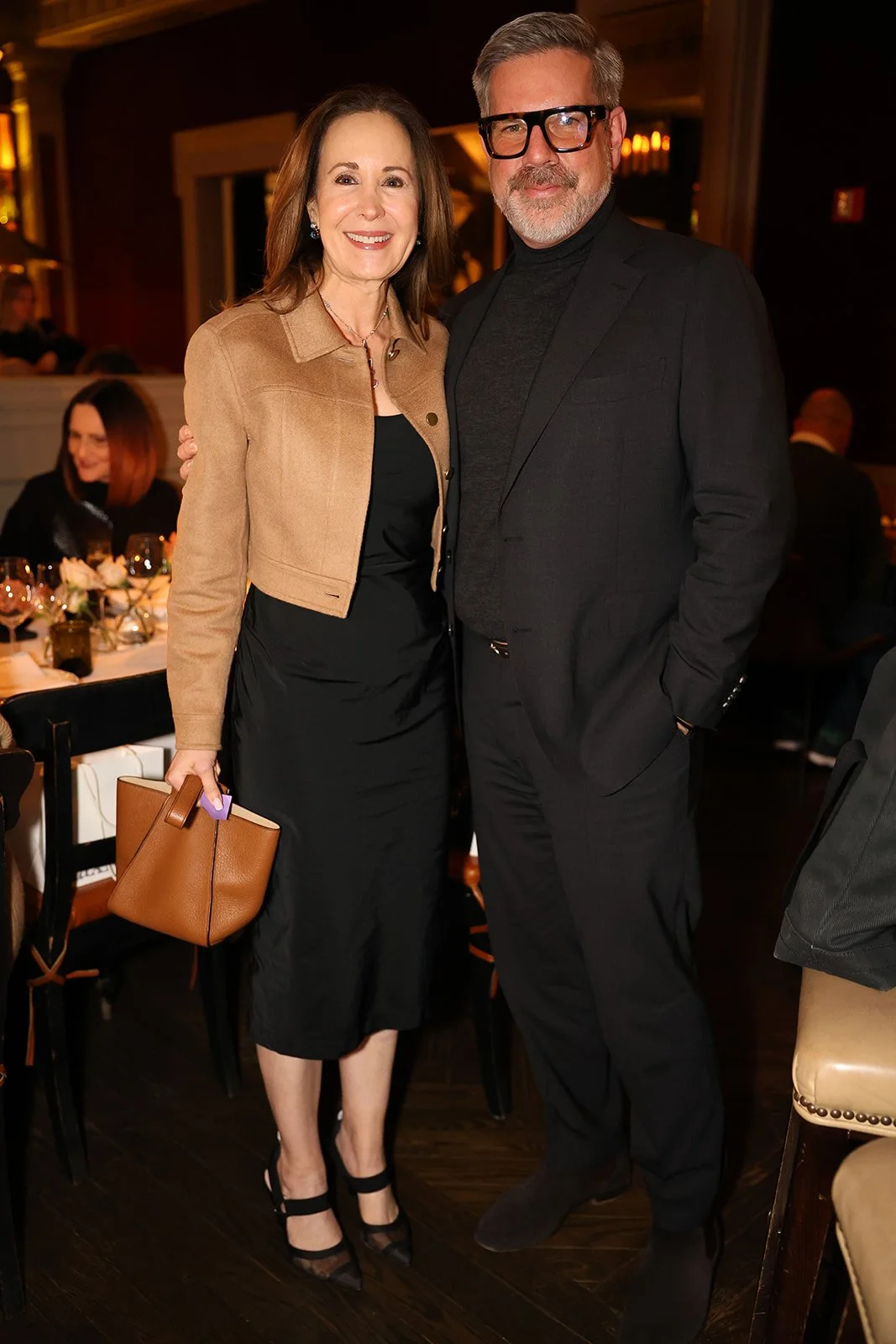 A smiling woman and a man are standing close together at an indoor event. The woman is holding a brown handbag and wearing a black dress with a light brown jacket, while the man is dressed in a black suit with glasses and a turtleneck. They are posin