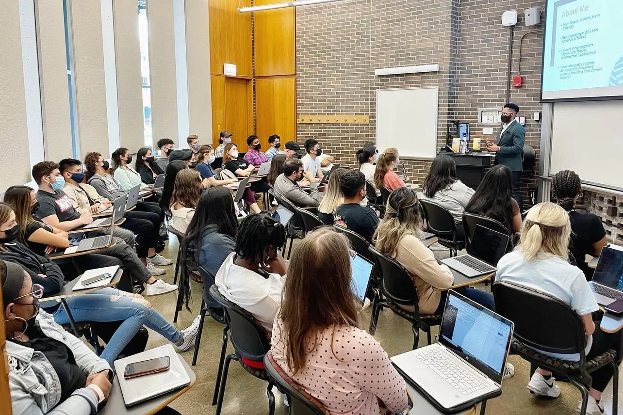 A classroom filled with students sitting at desks, many using laptops, listening to a male instructor at the front who is wearing a face mask, standing near a whiteboard and a large screen displaying a presentation.