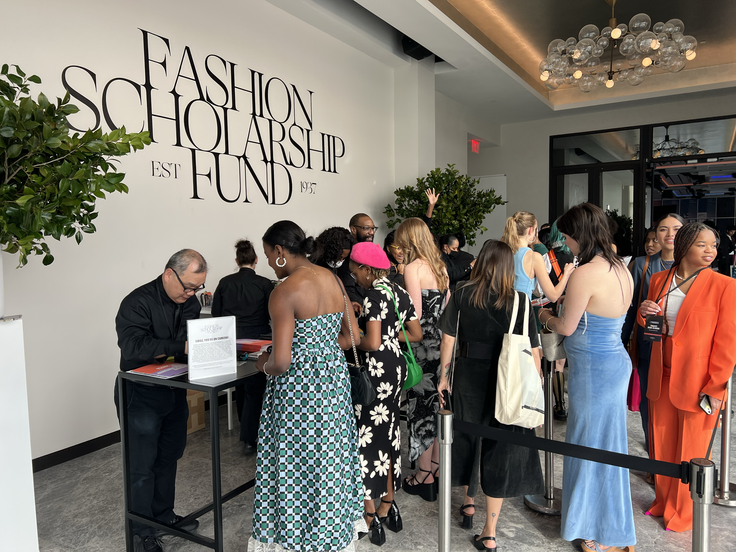 A diverse group of people in colorful outfits standing in line at a registration desk at an event called the Fashion Scholarship Fund, established in 1937, with a large sign on the wall and modern lighting fixtures overhead.