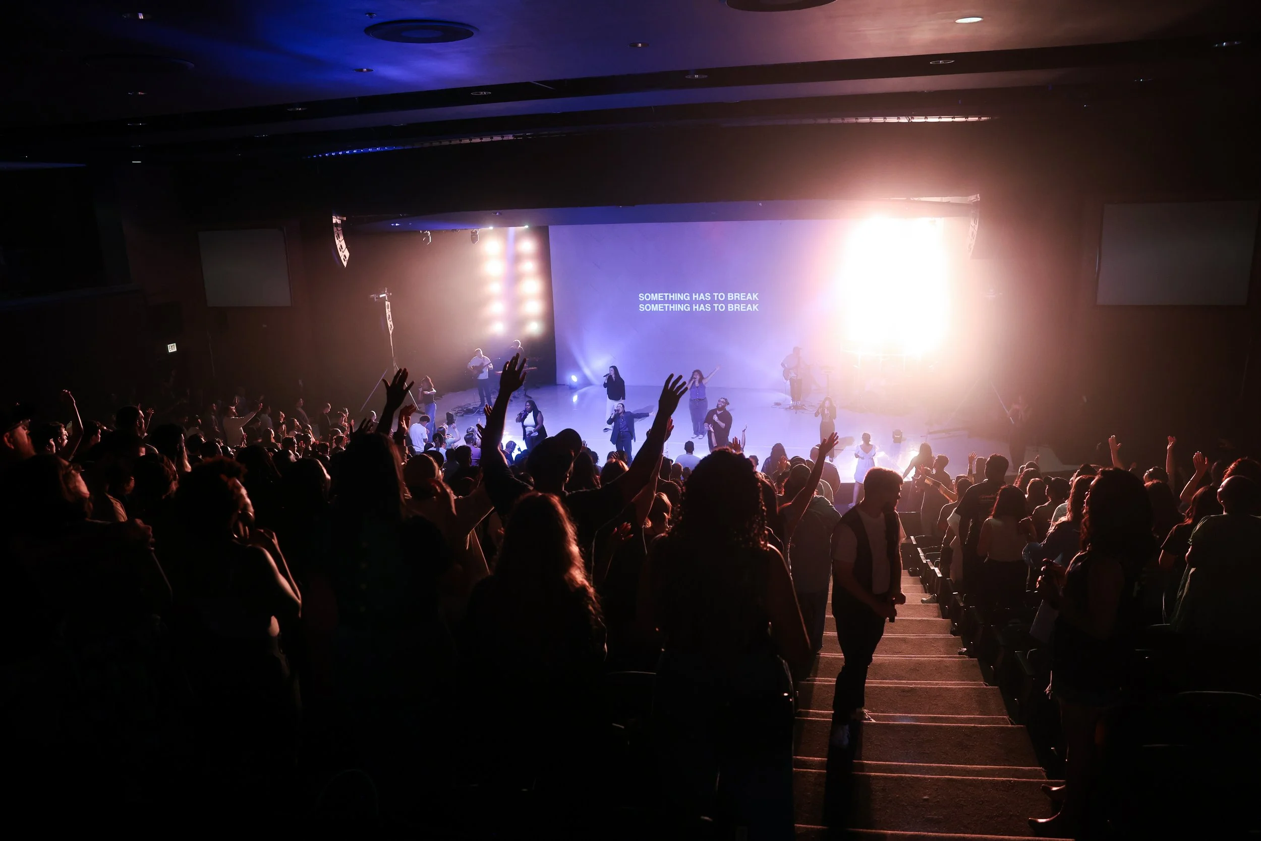 Crowd of people attending a concert or worship service with a stage, performers, and the lyrics 'SOMETHING HAS TO BREAK' projected on a large screen.