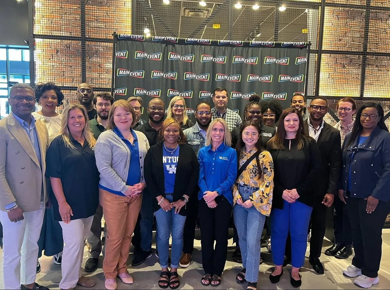 Group of diverse people posing for a photo in front of a black backdrop with 'Main Event' logos, inside a modern indoor space with metal and brick decor.
