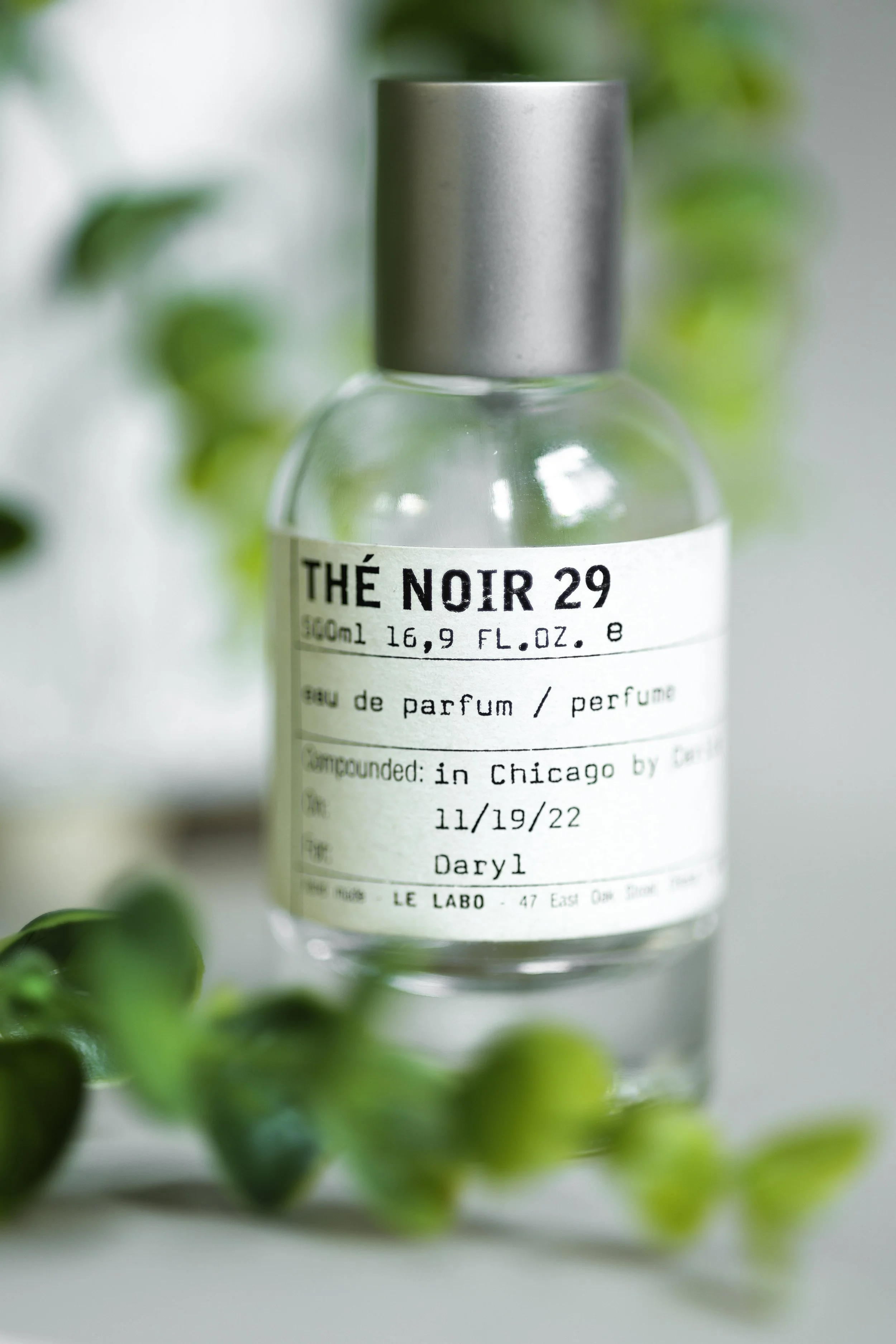 A clear glass perfume bottle labeled 'THÉ NOIR 29', with a metallic gray cap, surrounded by blurred green leaves.