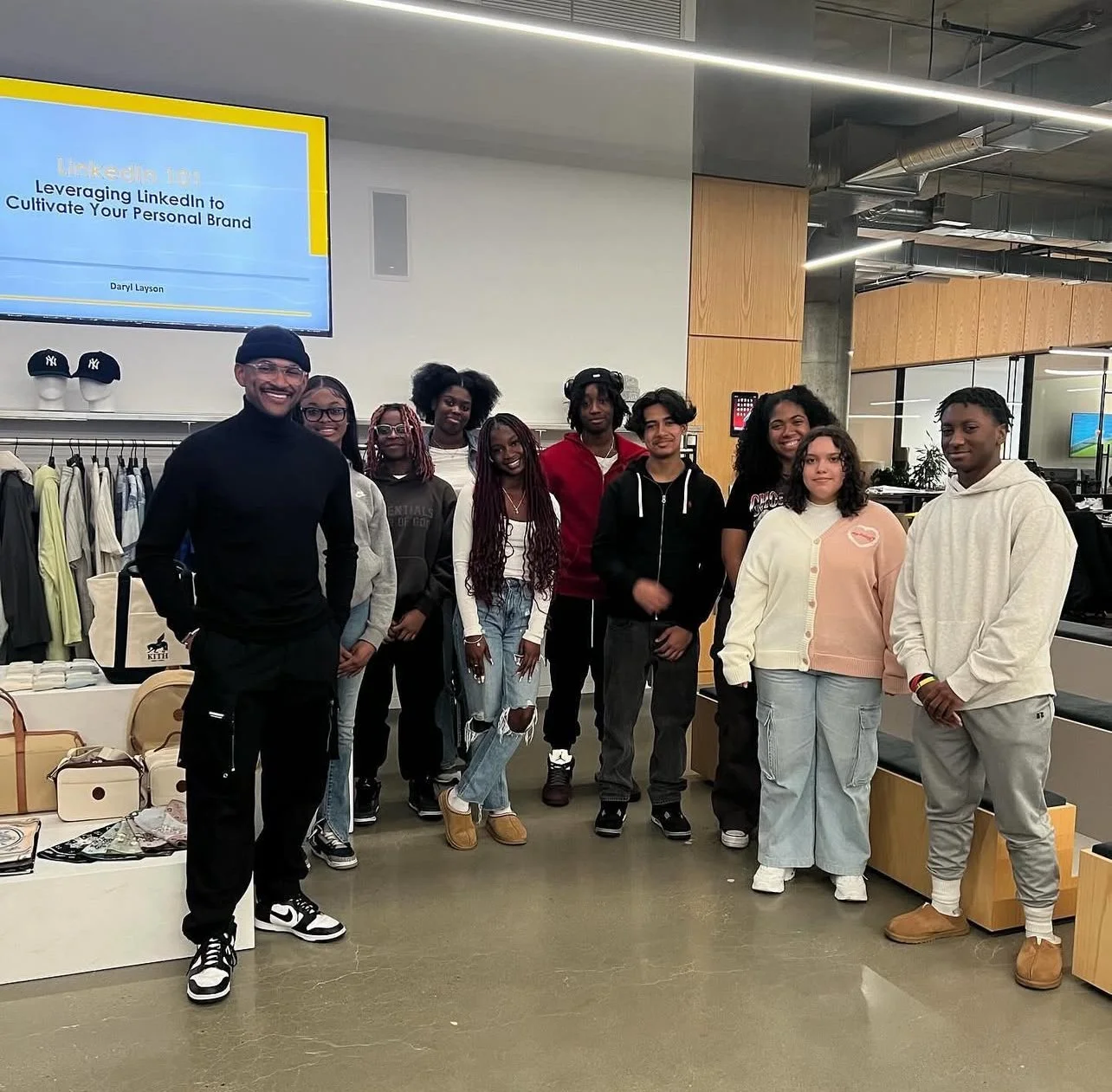Group of ten young people and adult man posing indoors in a retail store or office space, with clothing and accessories on display behind them.