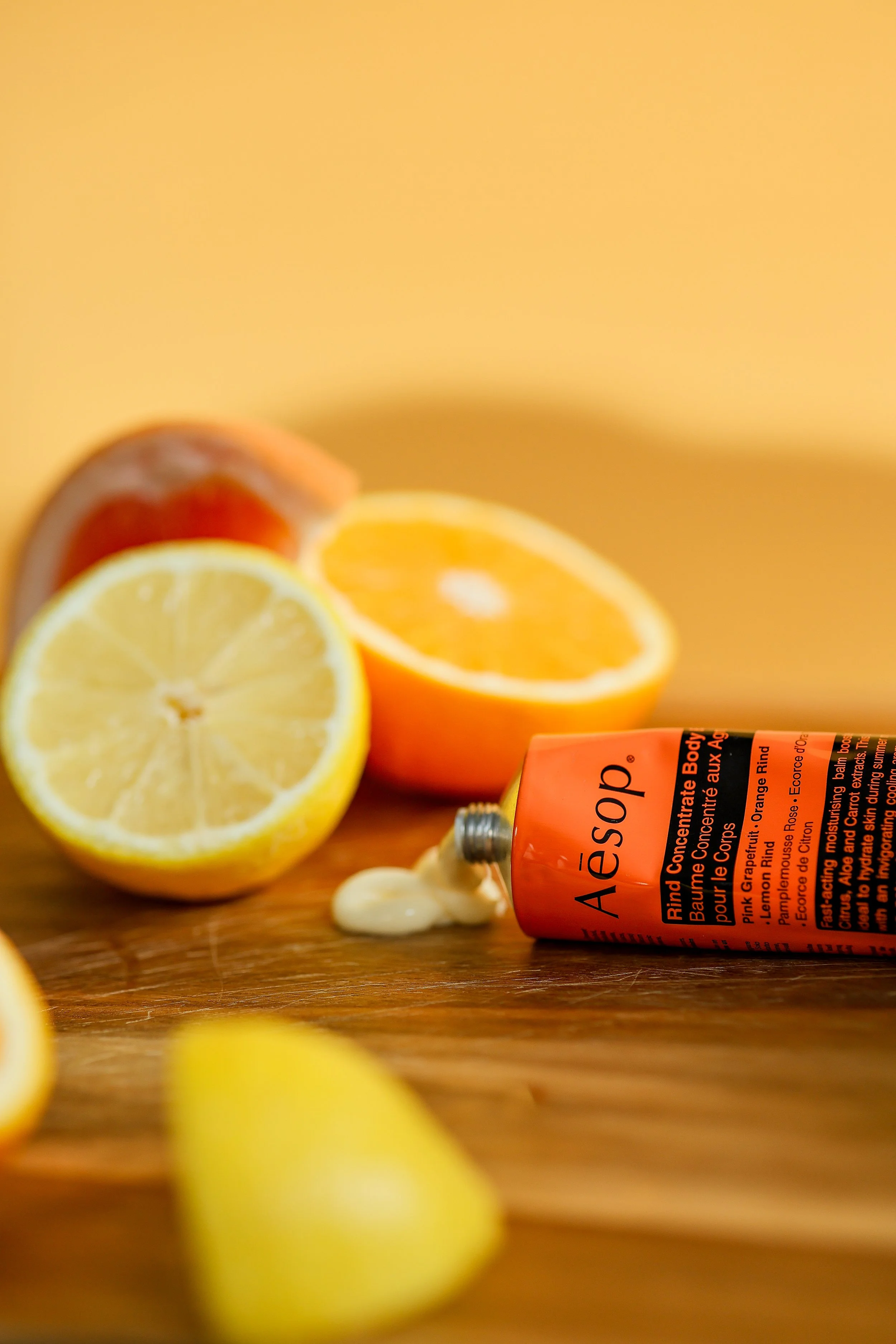 A tube of Aesop hand cream lying on its side next to sliced lemon and orange halves, with some cream visible on the wooden surface.