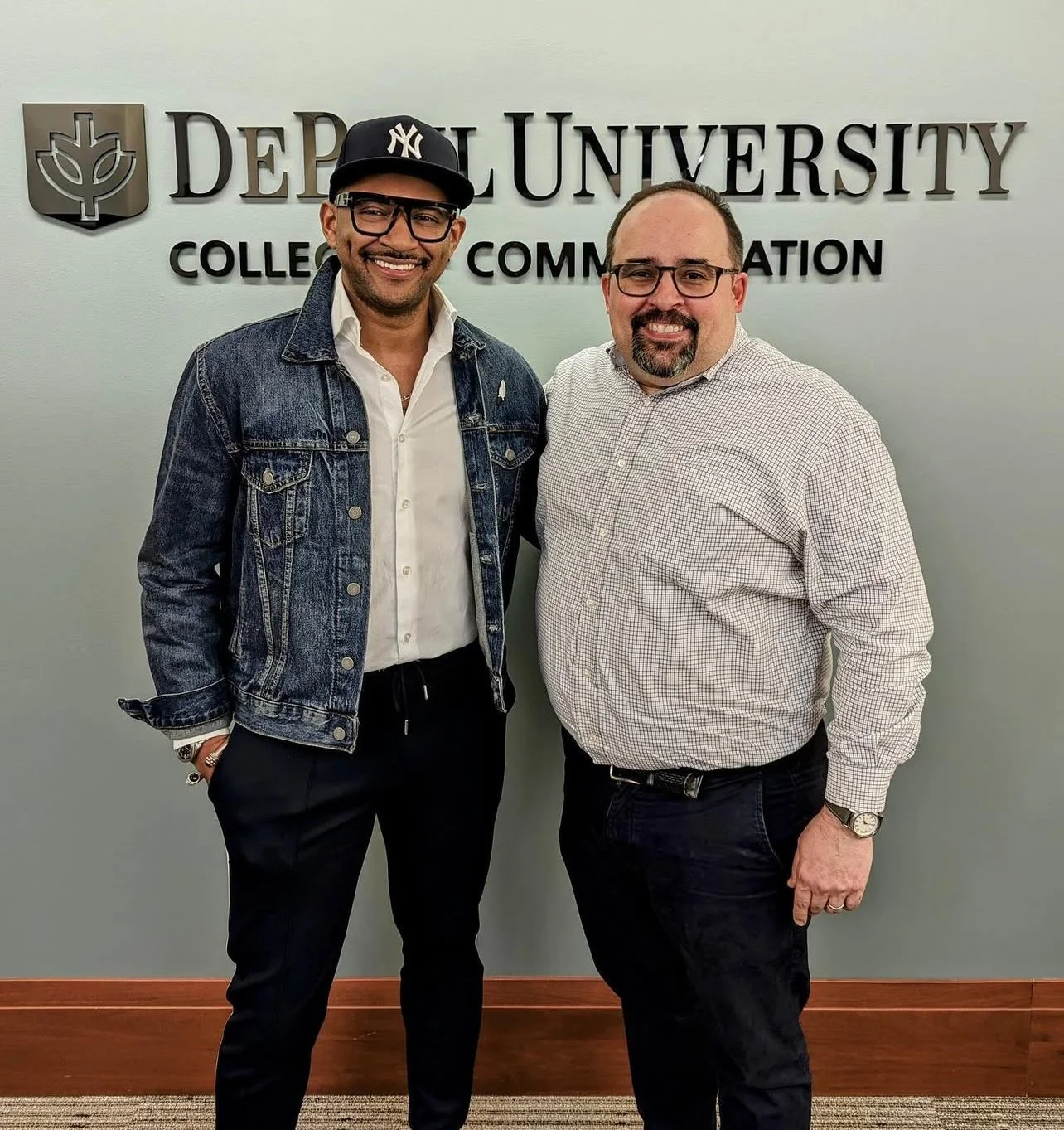 Two men smiling and standing together in front of a sign that reads DePaul University College of Communication. One man is wearing a black baseball cap, glasses, a denim jacket, a white shirt, and black pants. The other man is wearing glasses, a chec