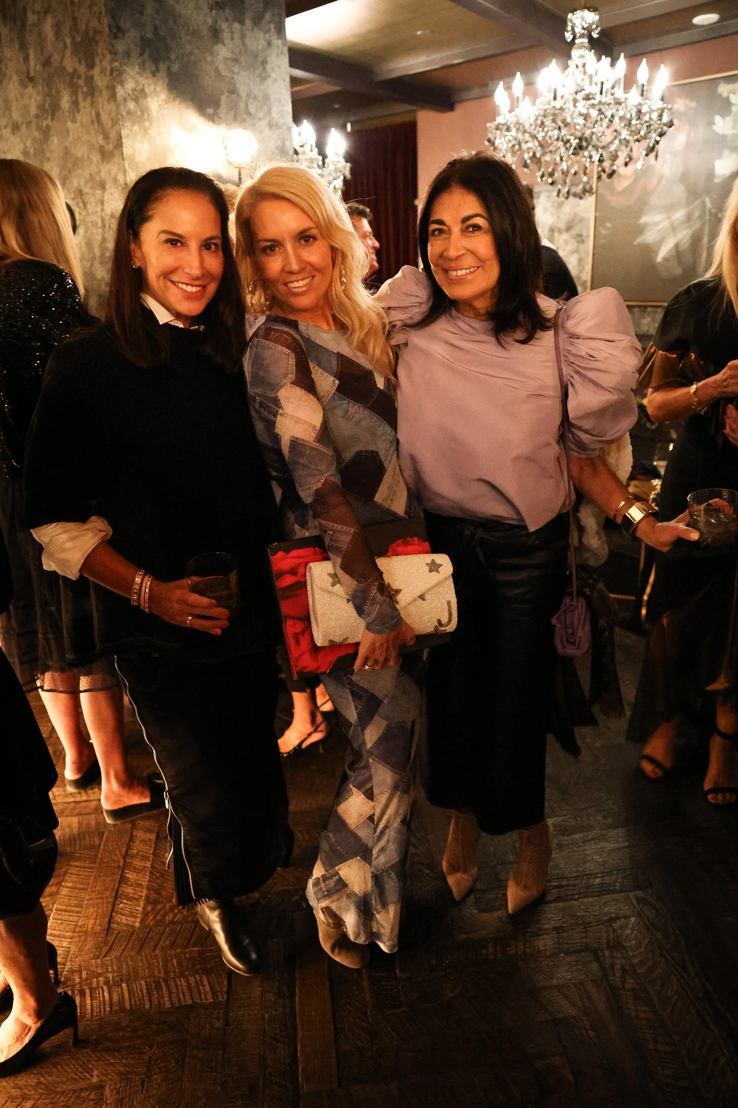 Three women smiling and posing together at a formal event, holding drinks, with chandeliers and other guests in the background.