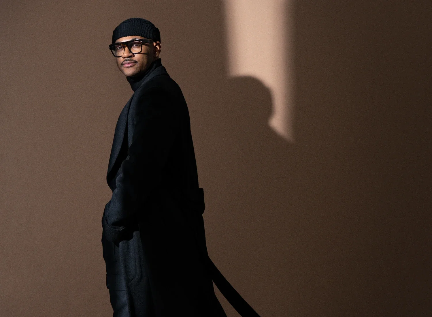 A man dressed in black, wearing glasses and a beanie, standing against a two-toned background with a shadow on the wall.