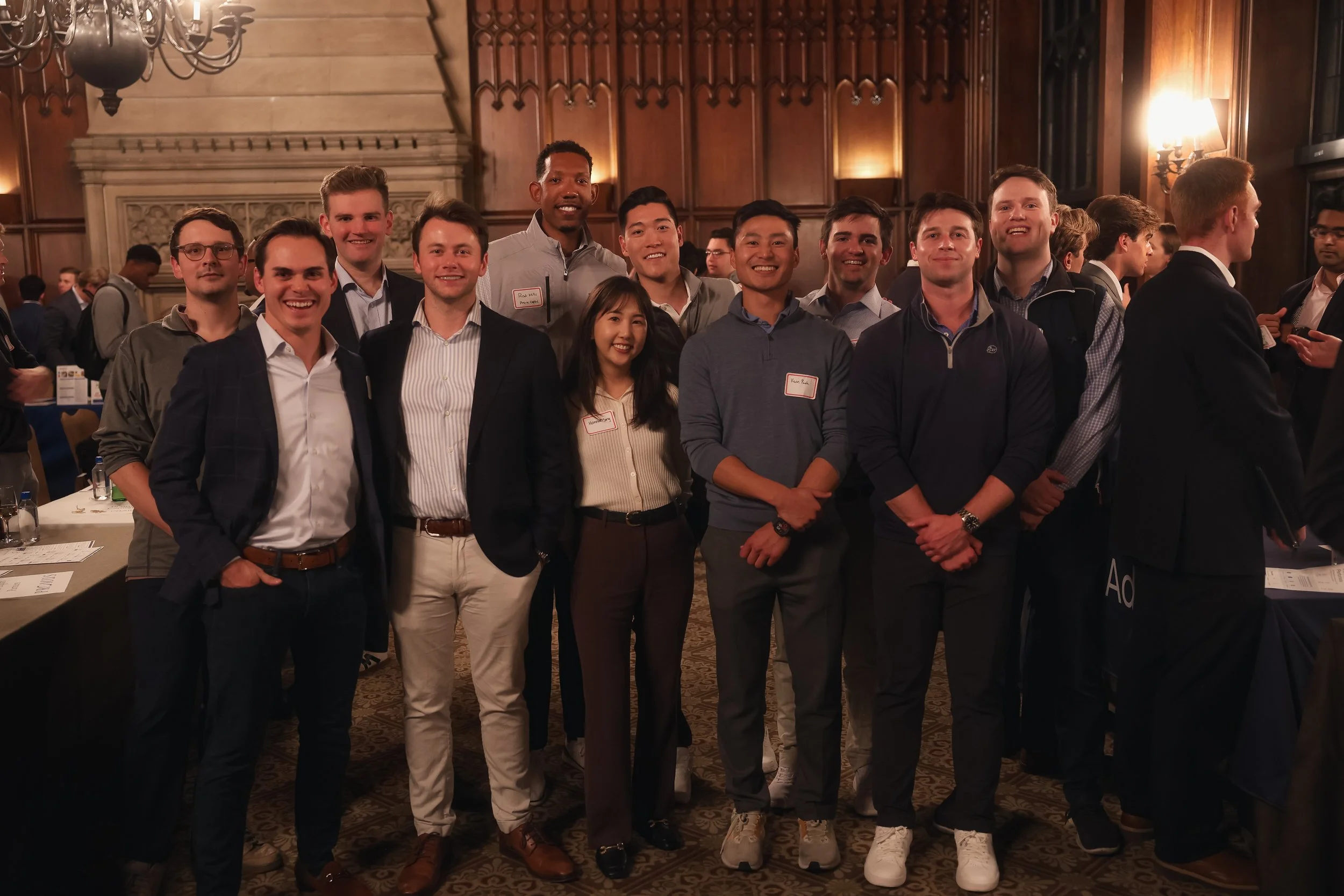 A group of young professionals, nine men and one woman, standing together at a formal indoor event in a grand, wood-paneled room with warm lighting, smiling at the camera.