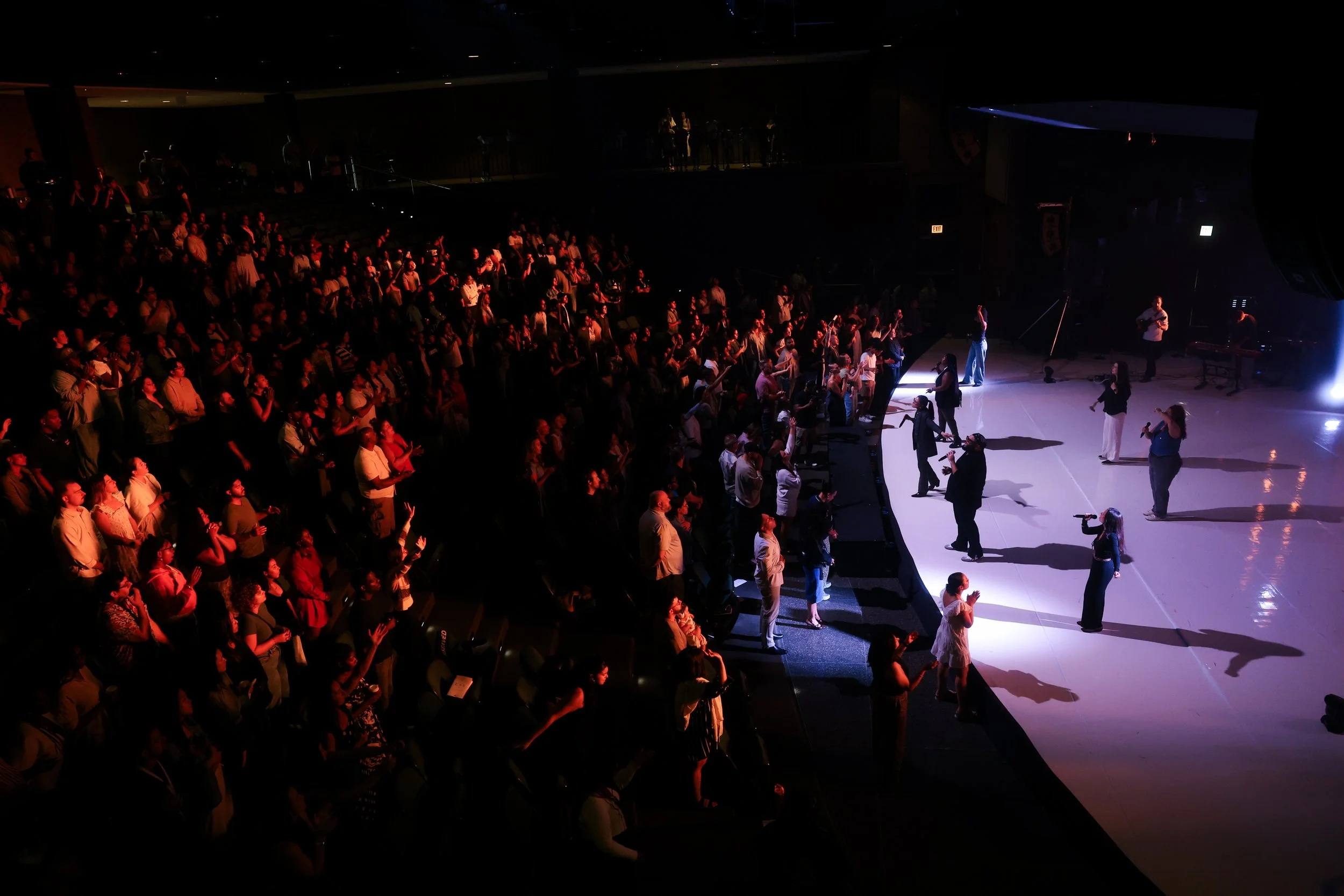 Audience gathered on a stage, some of whom are singing or performing, in a dark auditorium with dramatic lighting highlighting the performers.