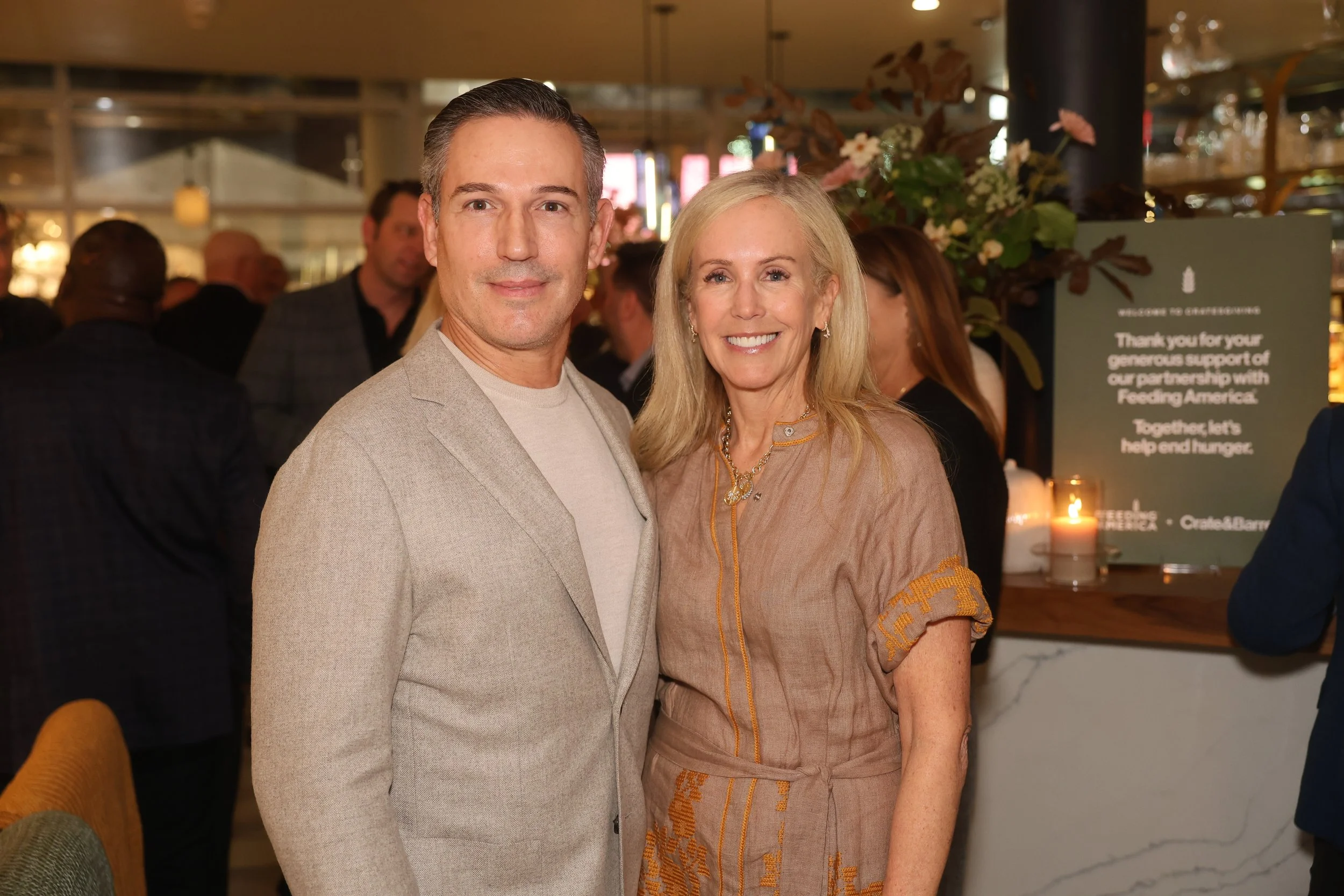 A man and woman standing close together at a gathering with other people in the background, smiling at the camera. The woman is wearing a tan dress with gold accents, and the man is wearing a light grey blazer over a beige shirt.