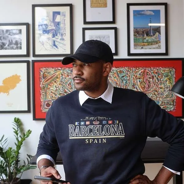 A man wearing a black cap and a dark blue sweater with 'Barcelona Spain' written on it sits in a room with framed artwork on the wall behind him, holding a phone.