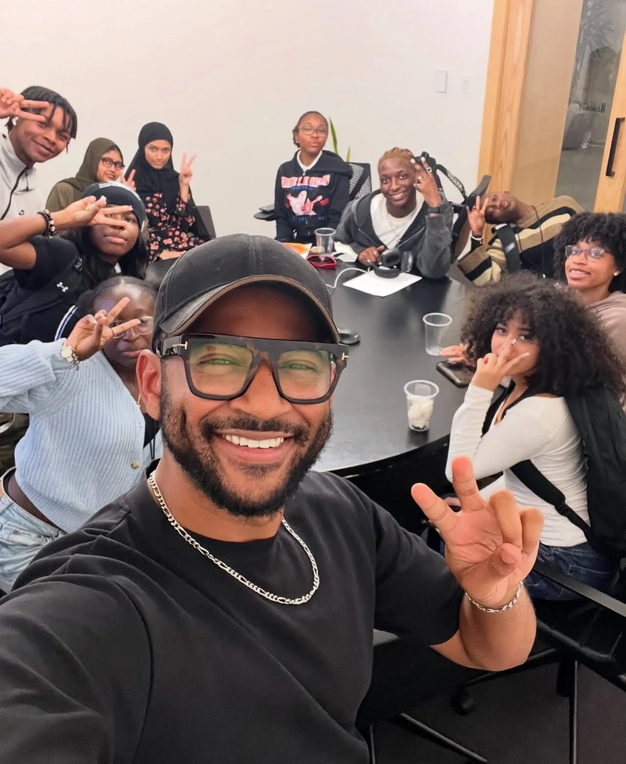 A group of diverse young people and one adult man taking a selfie at a round table in a room, all making peace signs or smiling.