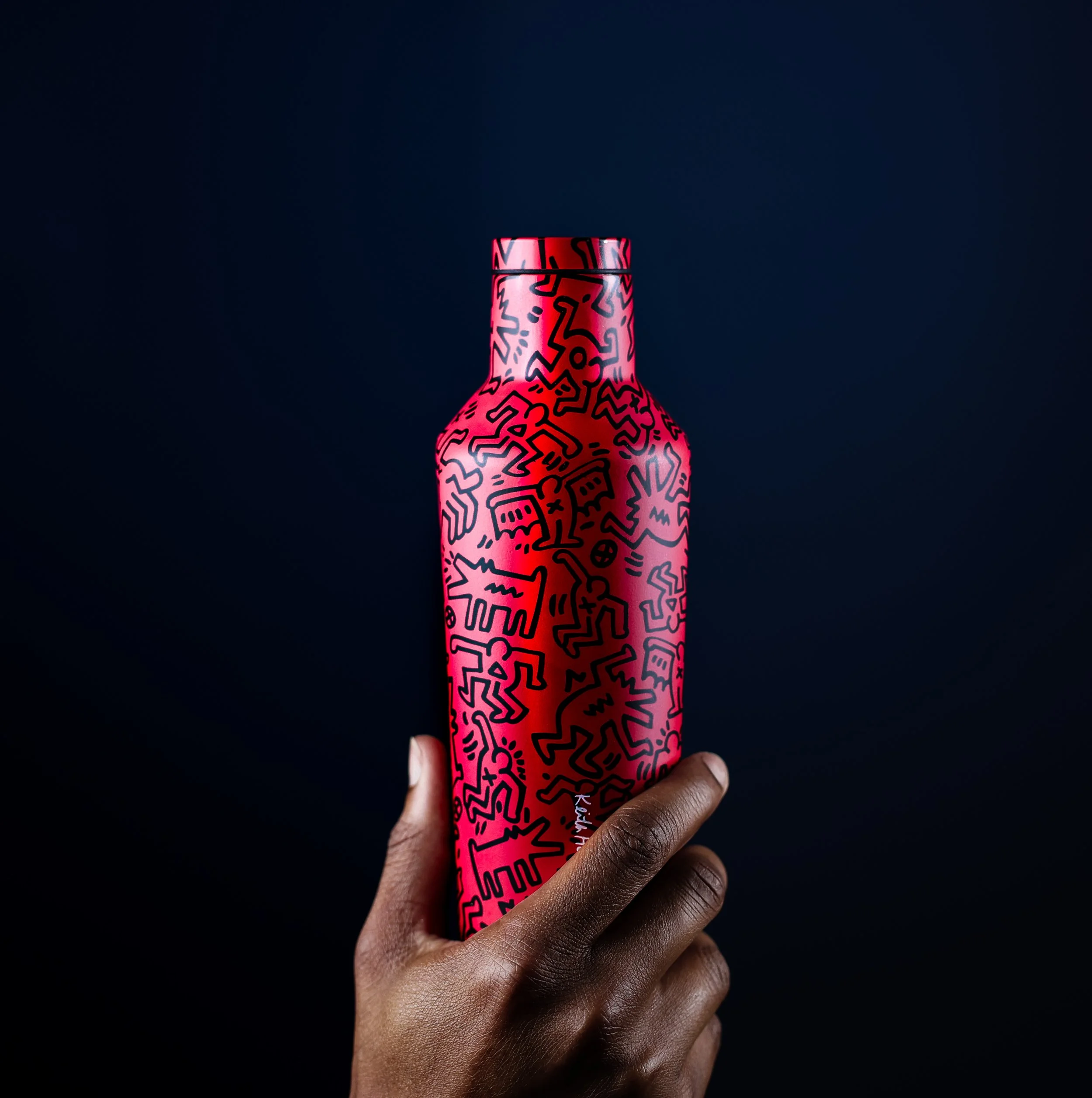 A hand holding a red bottle with abstract black line art designs on it, against a dark background.