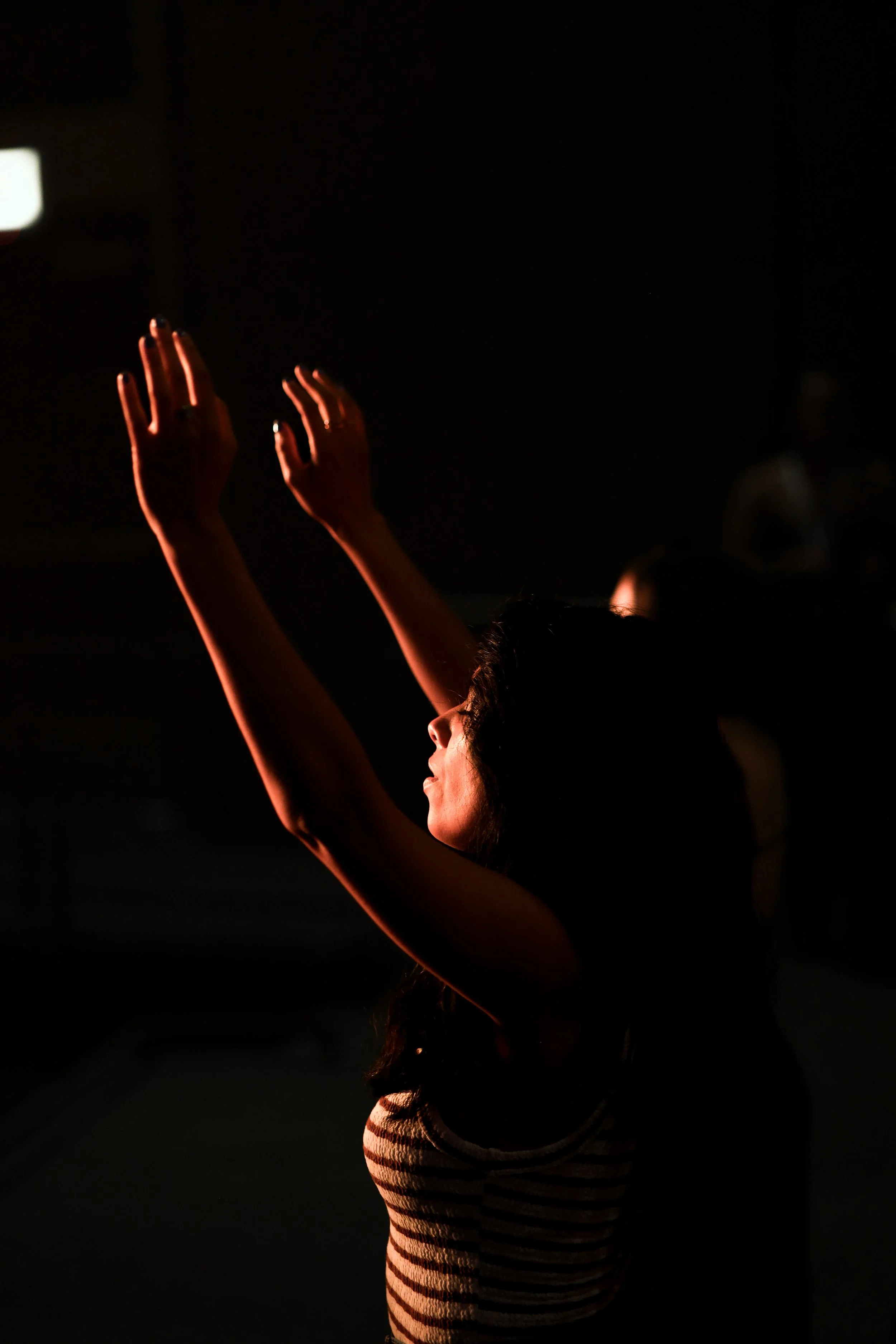 A woman with dark hair and a striped shirt raises her arms with her face turned slightly upward, illuminated by a warm light in a dark setting.