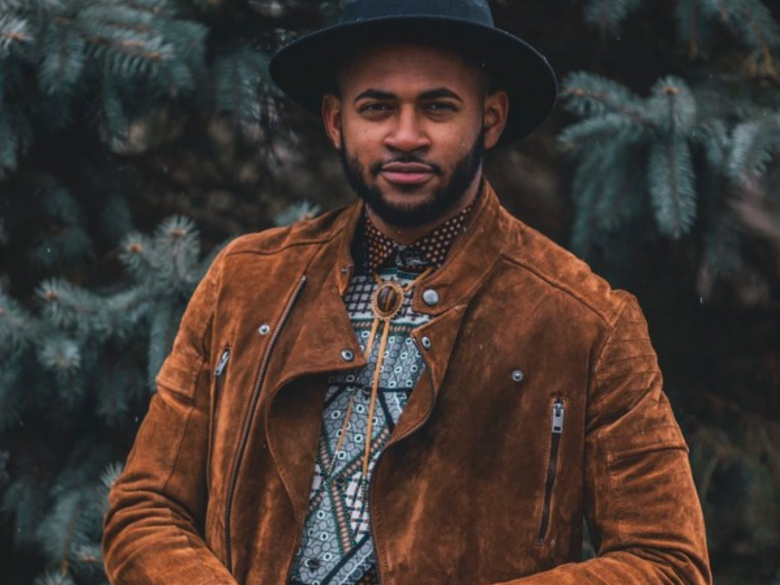A man with a beard wearing a black hat, brown suede jacket, and patterned shirt standing in front of pine trees.