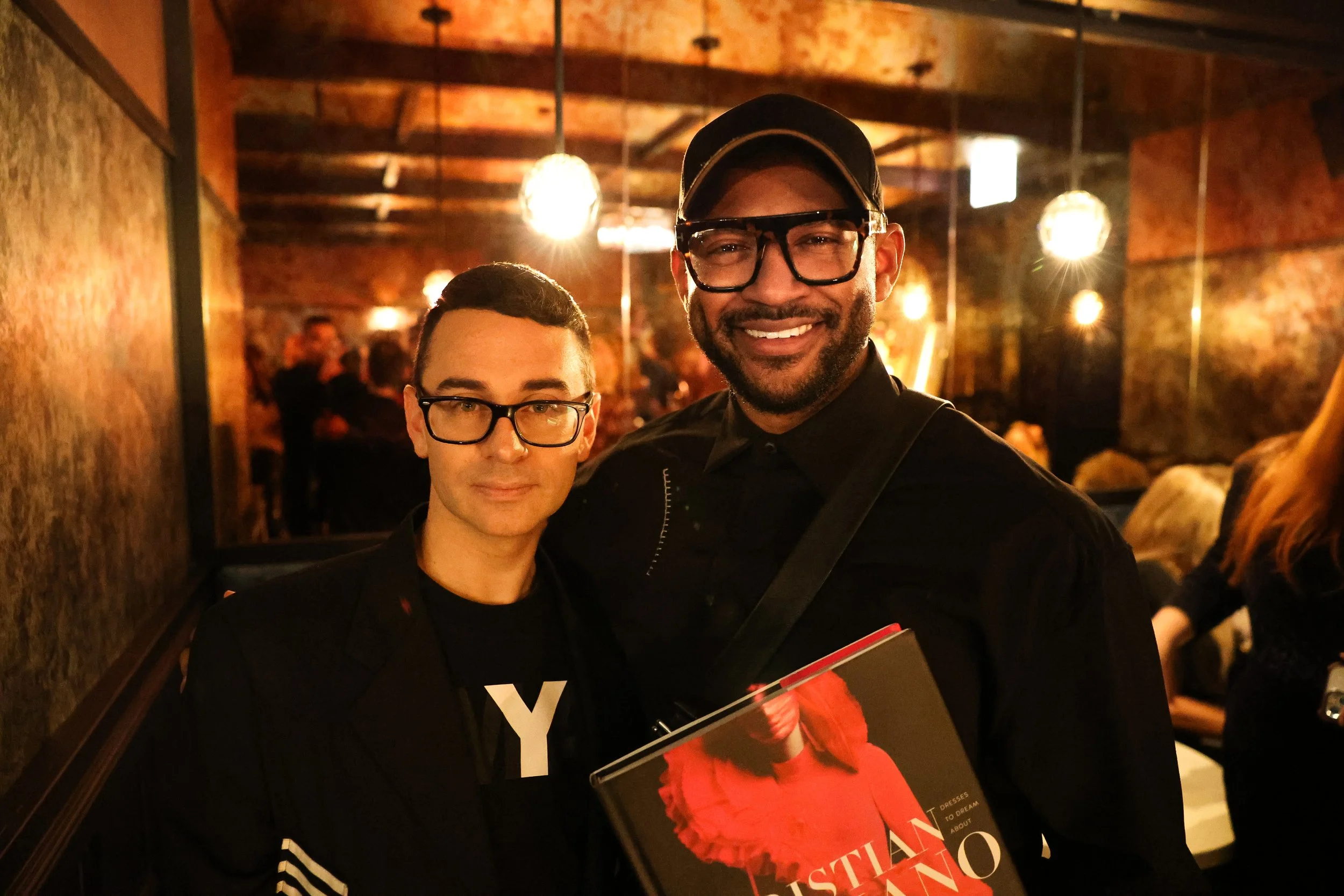 Two men with glasses smiling, one holding a book, standing in a dimly lit, rustic indoor setting with warm lighting and exposed brick walls.