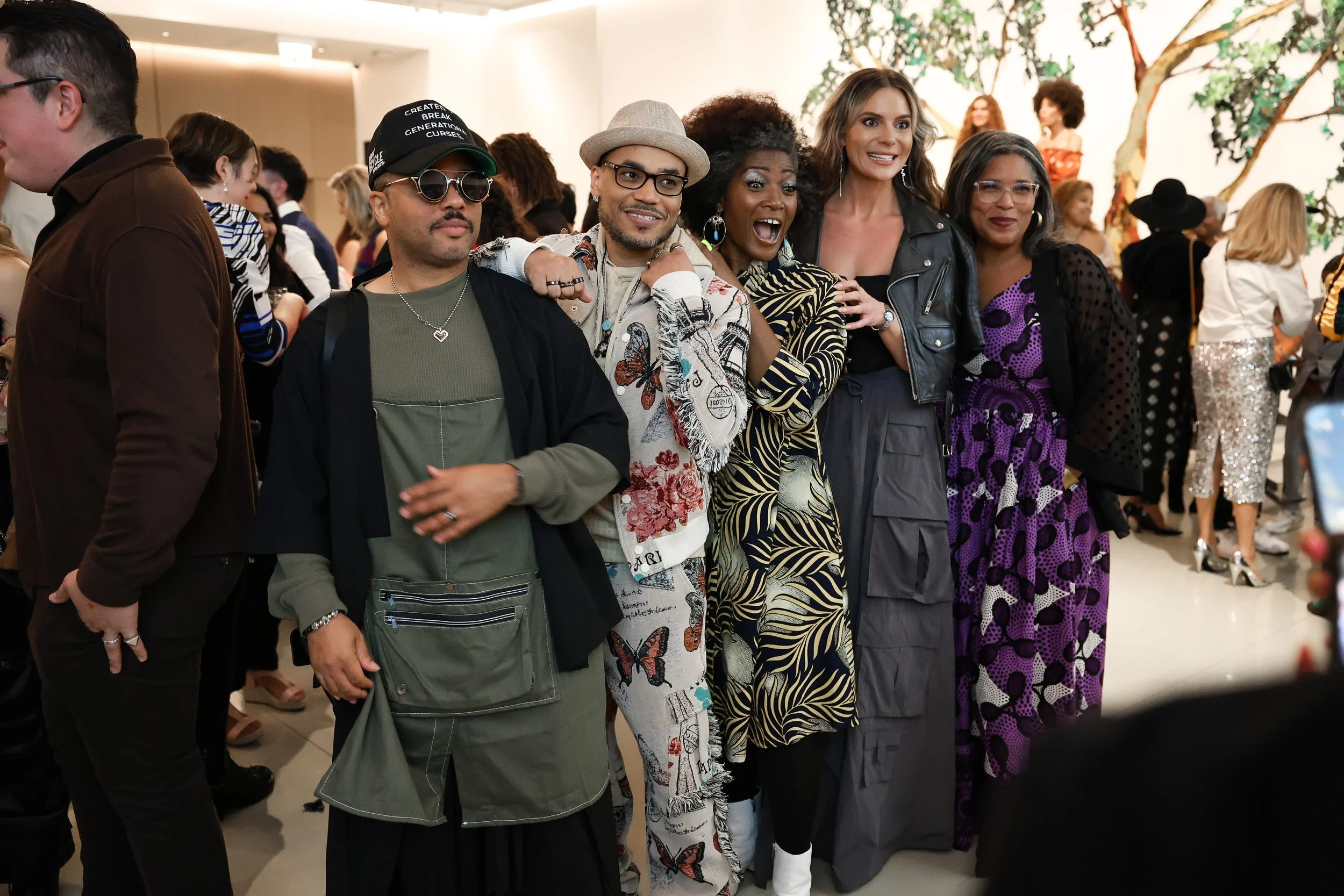Group of five people standing together, smiling and posing, at an indoor social event with other people in the background.
