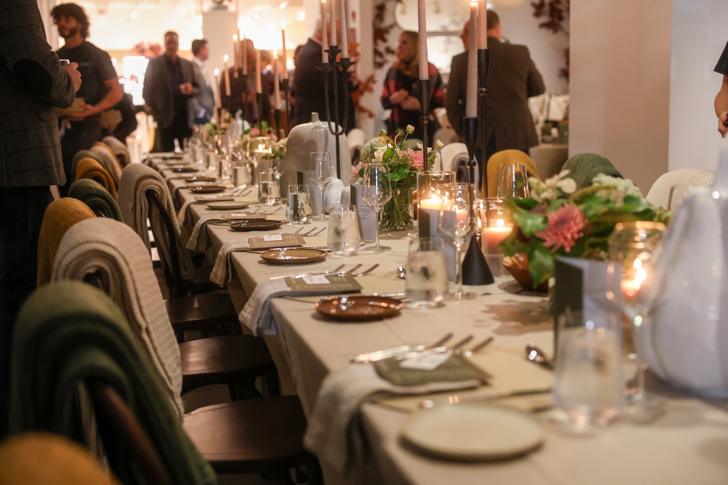 A long dining table set for a formal event with candles, flowers, plates, glasses, and cutlery, in a warmly lit room with people standing and conversing in the background.