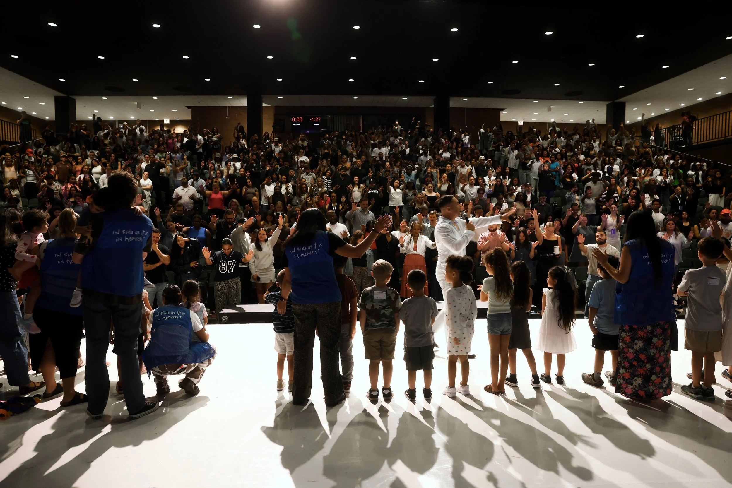 A large crowd gathered in an auditorium or indoor venue, with some people standing on stage or platform in front, participating in a group event or worship service. Children and adults are seen raising hands, singing, and engaging in the activity.