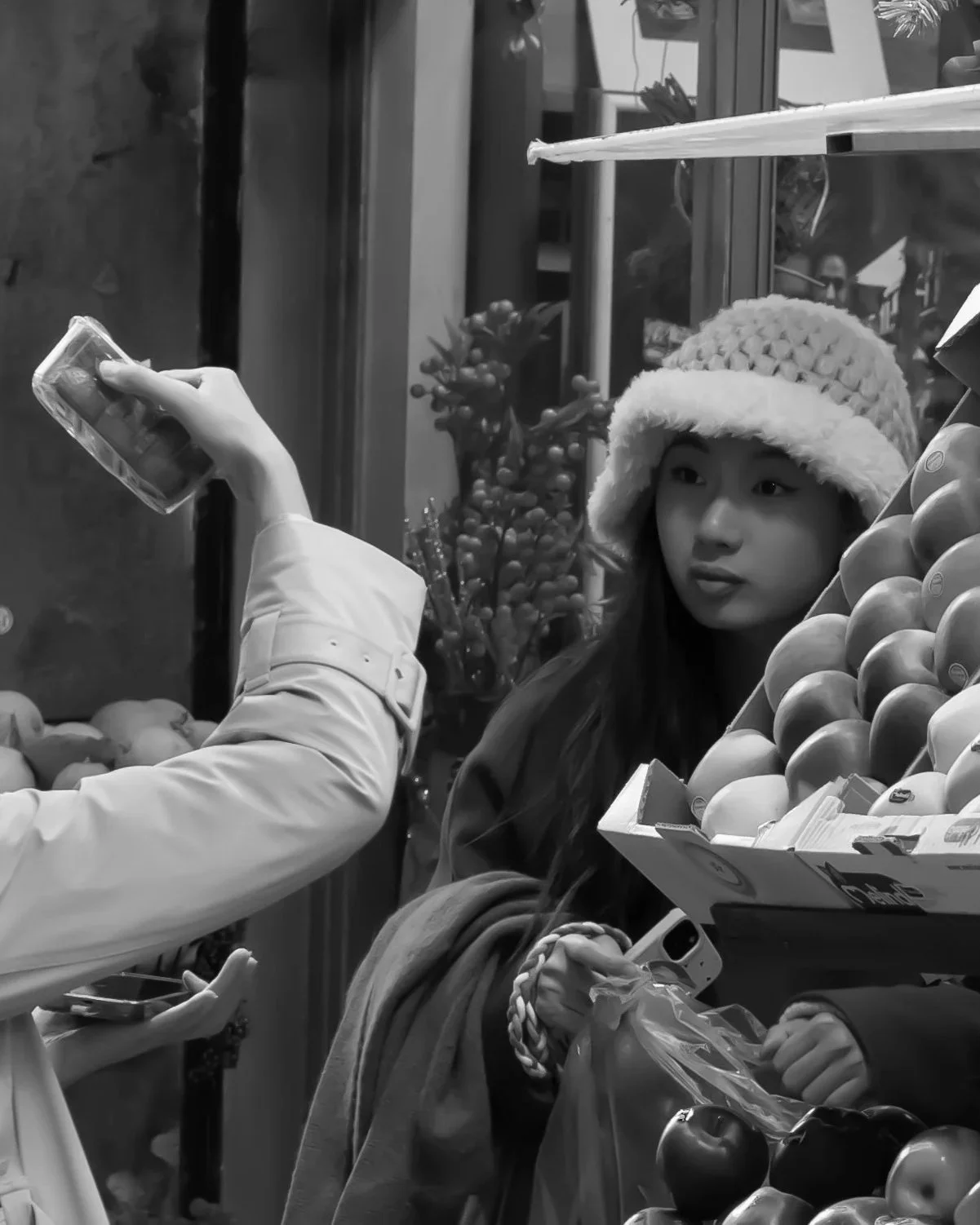 A woman with light skin and dark hair, wearing a cozy hat, is at a produce stand with apples. Someone else is taking a photo or video of her with a smartphone. The scene appears to be outdoors with various fruits and plants in the background.