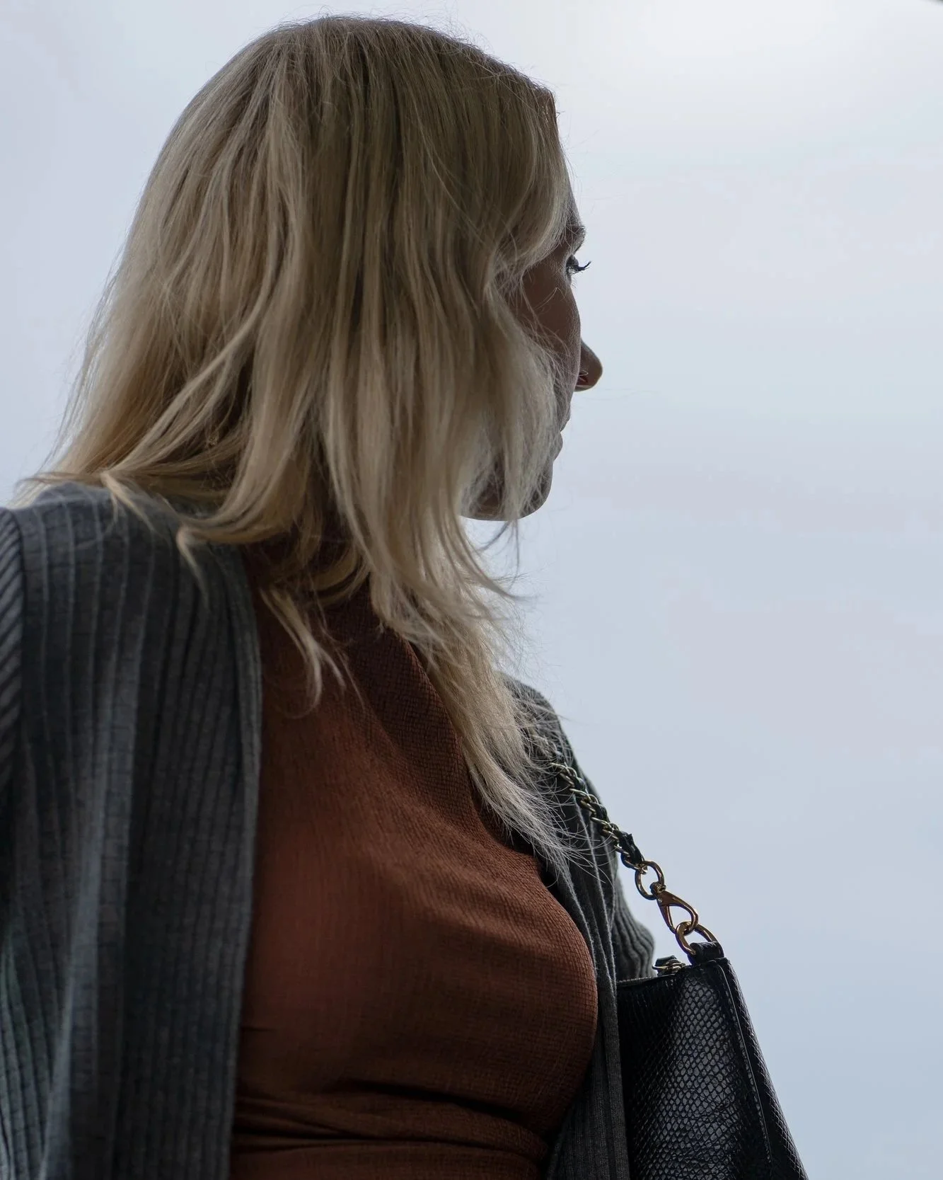 A woman with blonde hair wearing a brown top and a striped blazer, carrying a black purse with a chain strap, looking to the right against a cloudy sky.