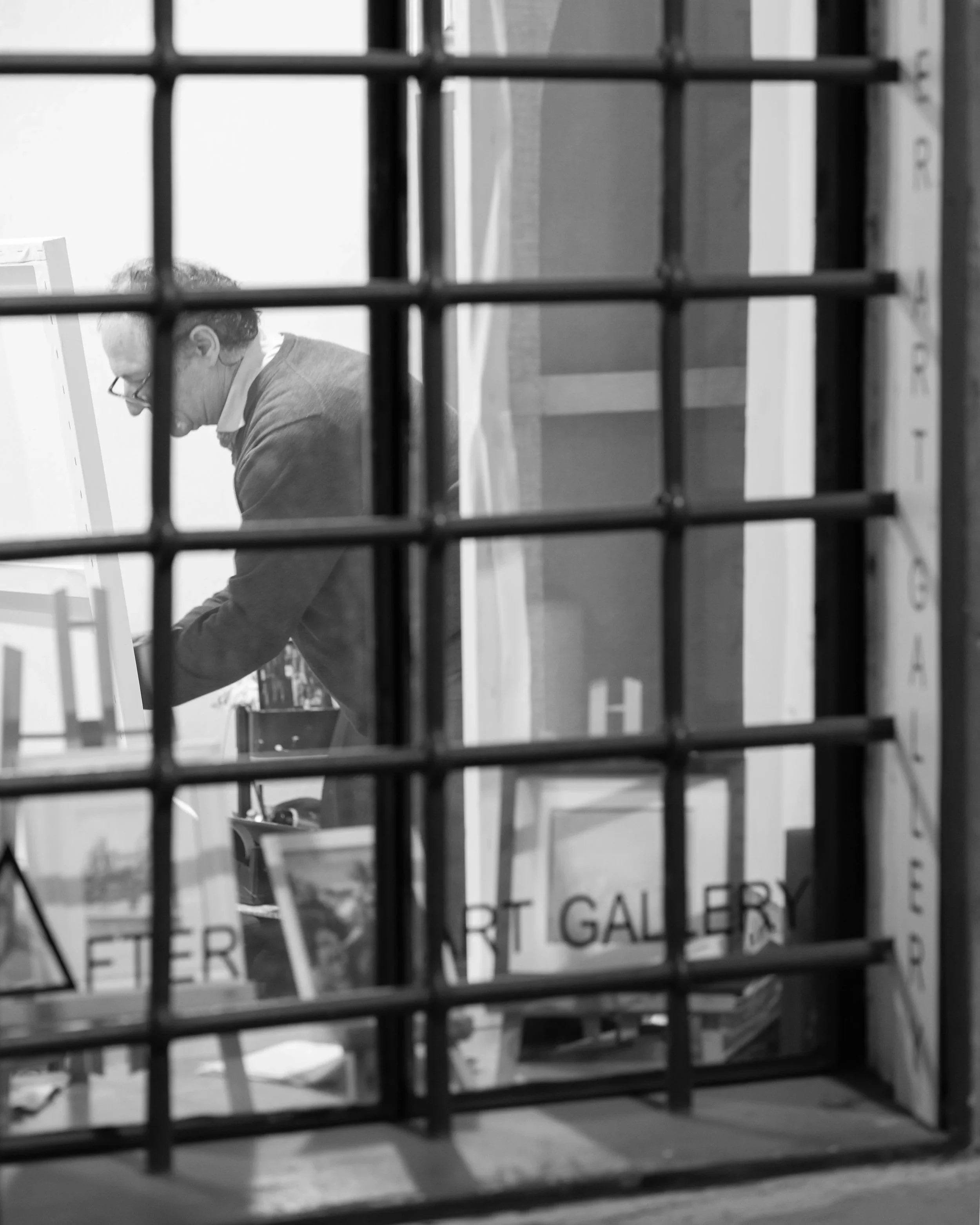 A person with glasses working at a desk is viewed through a grid-like barrier with square openings, creating a layered, framed perspective.