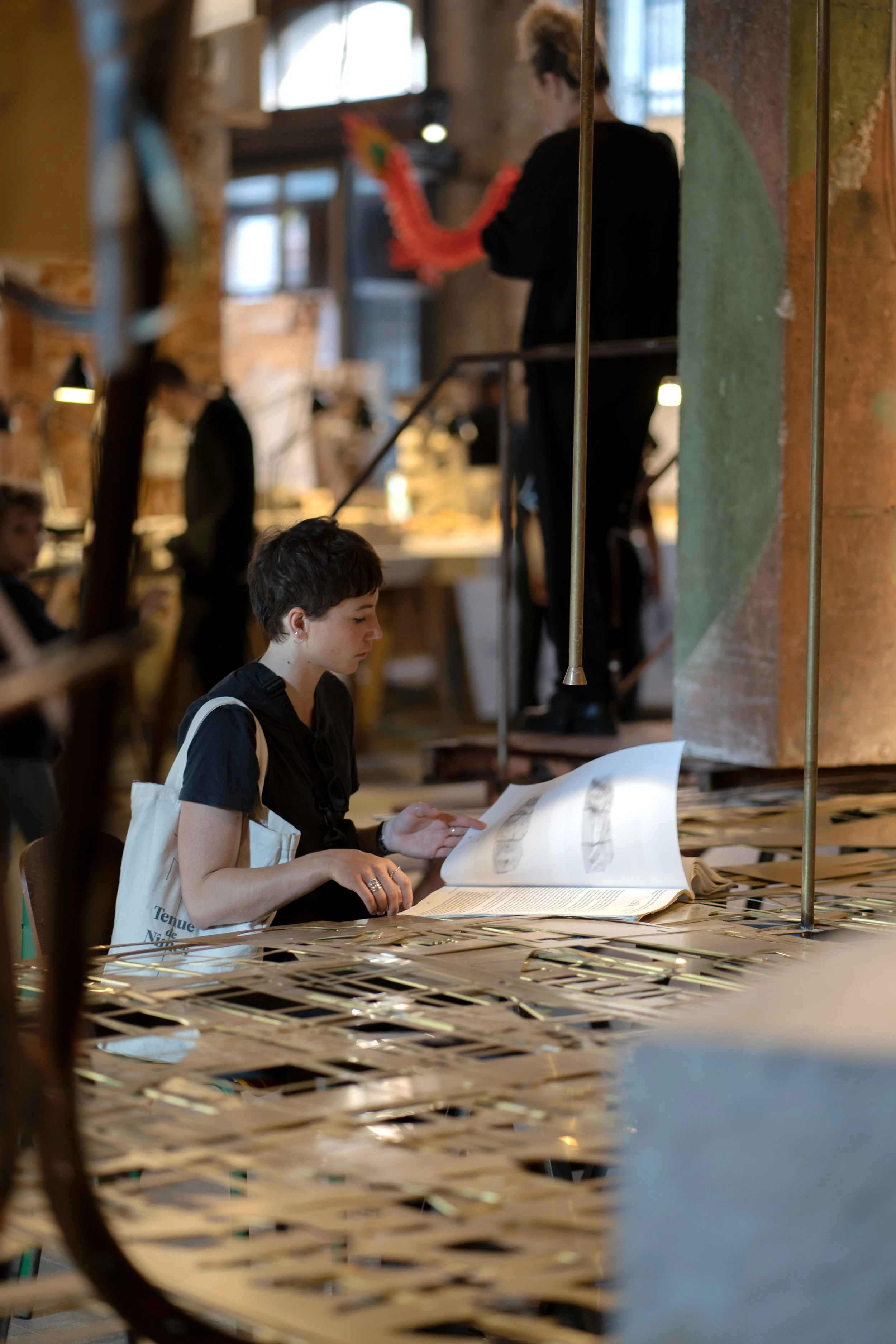 A young woman with short dark hair sits at a metallic table in a busy, industrial-style cafe or art space, reading a large book. In the background, a person on a staircase appears to be preparing or displaying colorful artwork, with other people engaged in different activities.