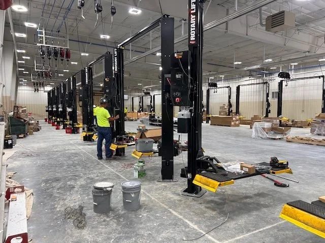Workers installing or maintaining large black machine frames with yellow bases in an industrial warehouse setting.