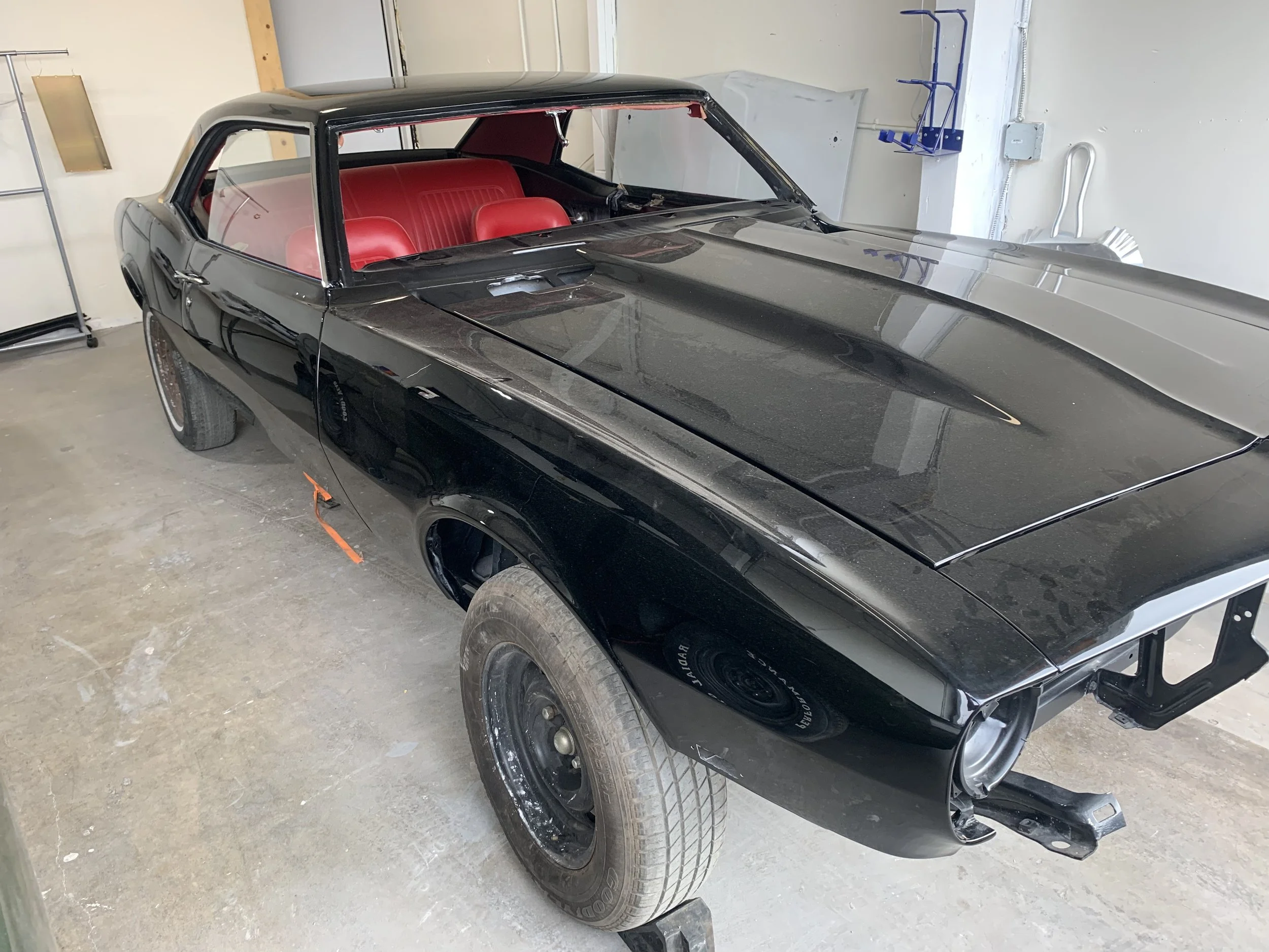 Black vintage muscle car with red interior in a garage. Front wheel missing, car appears to be under restoration.