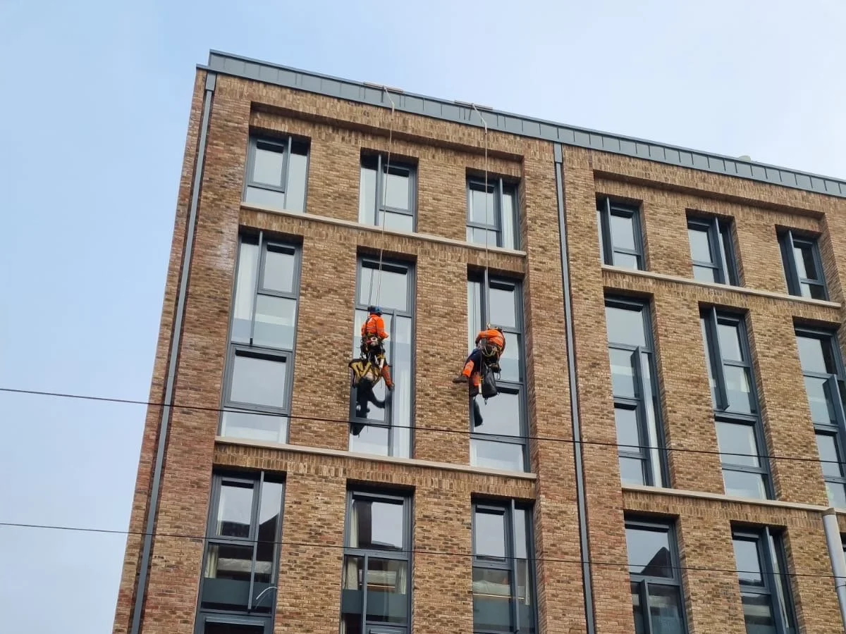 Two window washers in orange safety gear cleaning windows on a brick building.