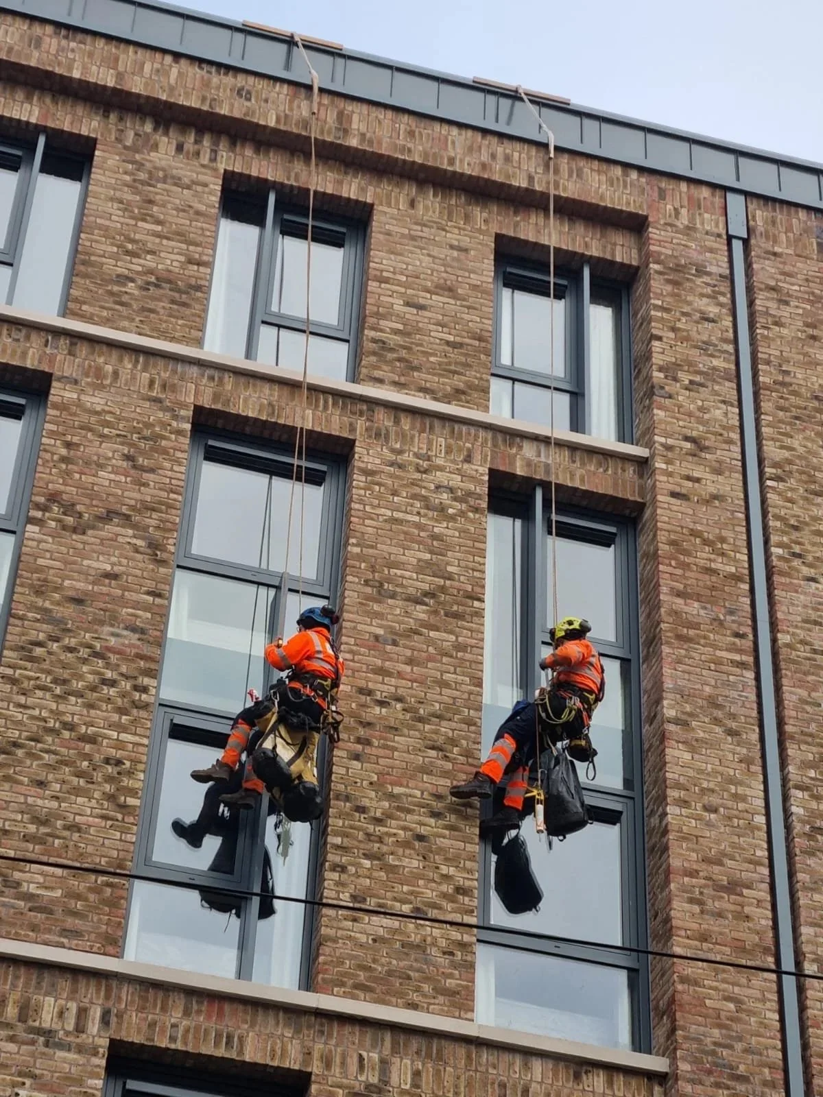 Two window cleaners wearing orange uniforms and safety helmets are hanging on harnesses outside a multi-story brick building, cleaning the windows.